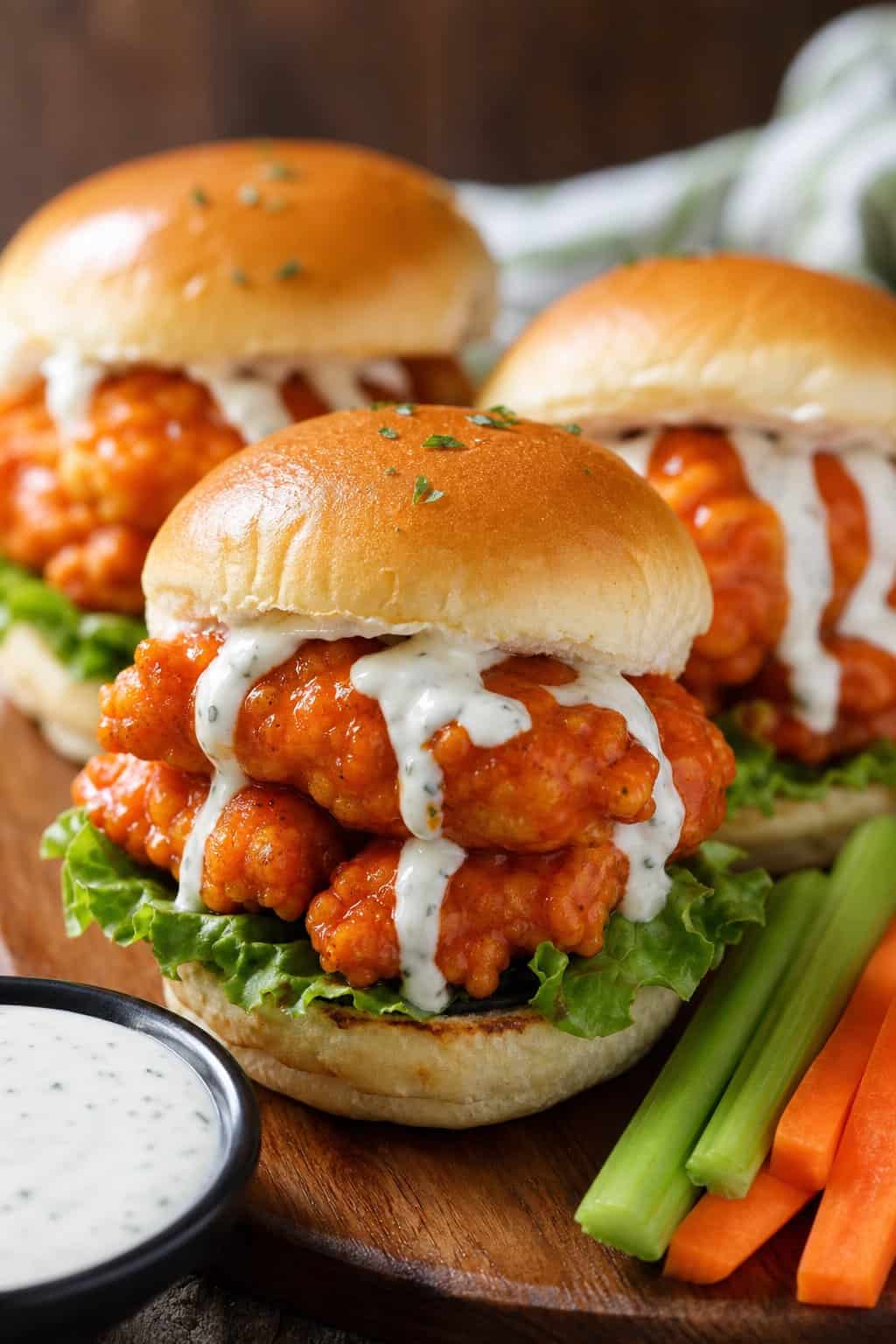 Close-up of buffalo ranch chicken wing sliders with lettuce on a wooden board, served with celery, carrot sticks, and a bowl of ranch dressing.