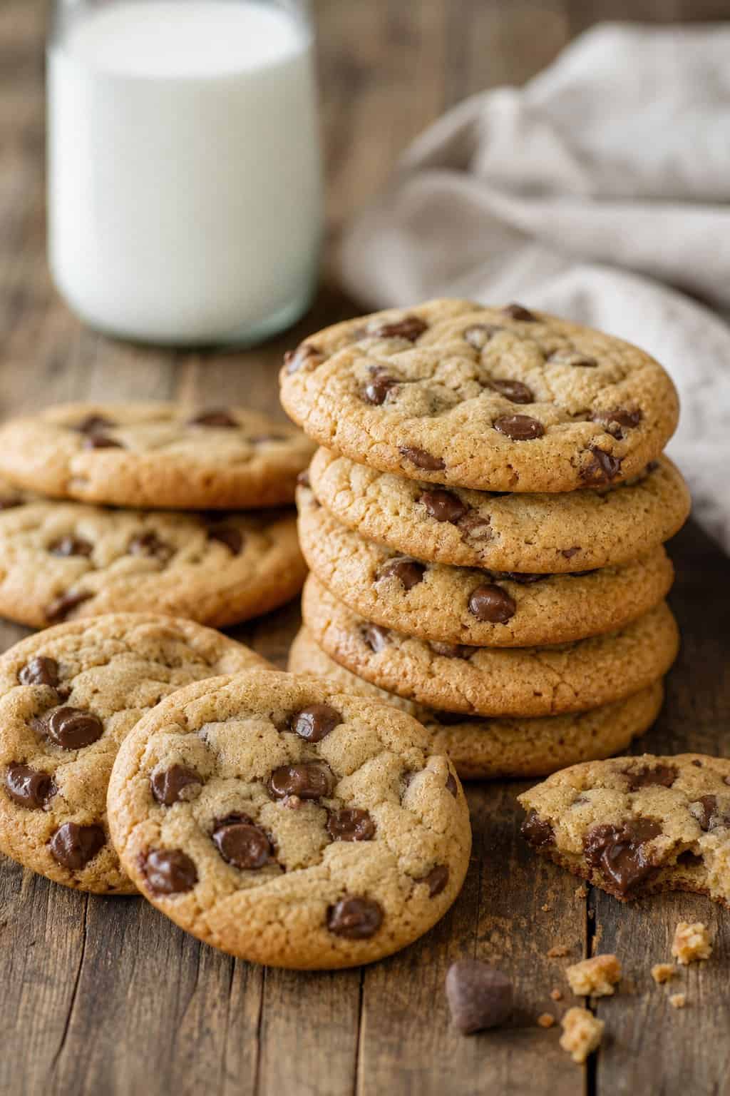 A close-up of freshly baked chocolate chip cookies on a wooden table with a glass of milk in the background.