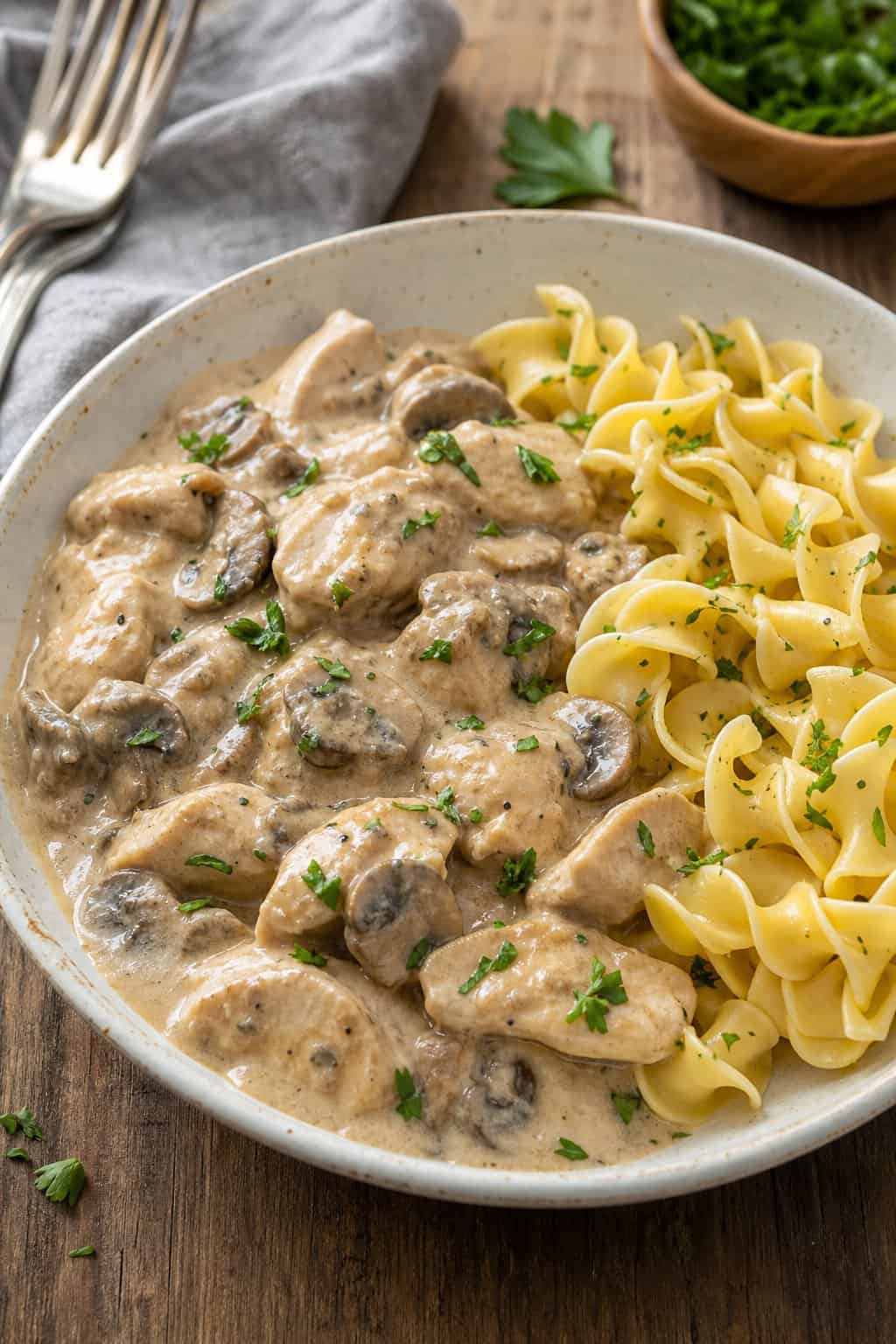 A bowl of creamy mushroom chicken stroganoff with egg noodles and parsley garnish on a wooden table.