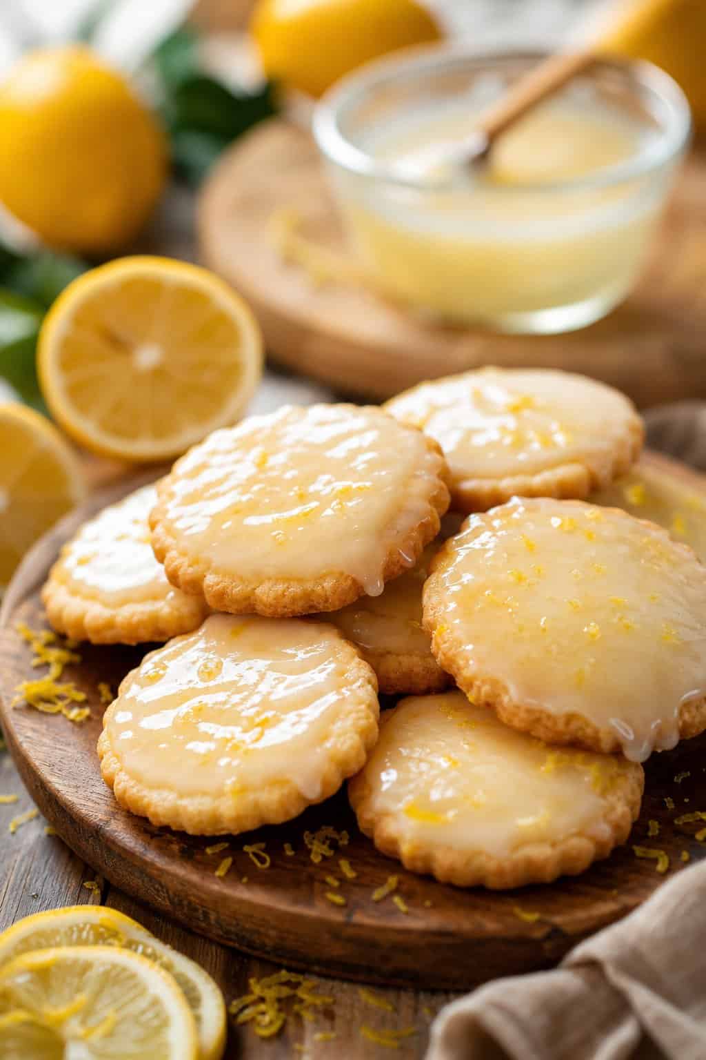 A close-up of lemon-glaze shortbread cookies on a wooden platter with fresh lemon slices and a bowl of lemon glaze nearby.