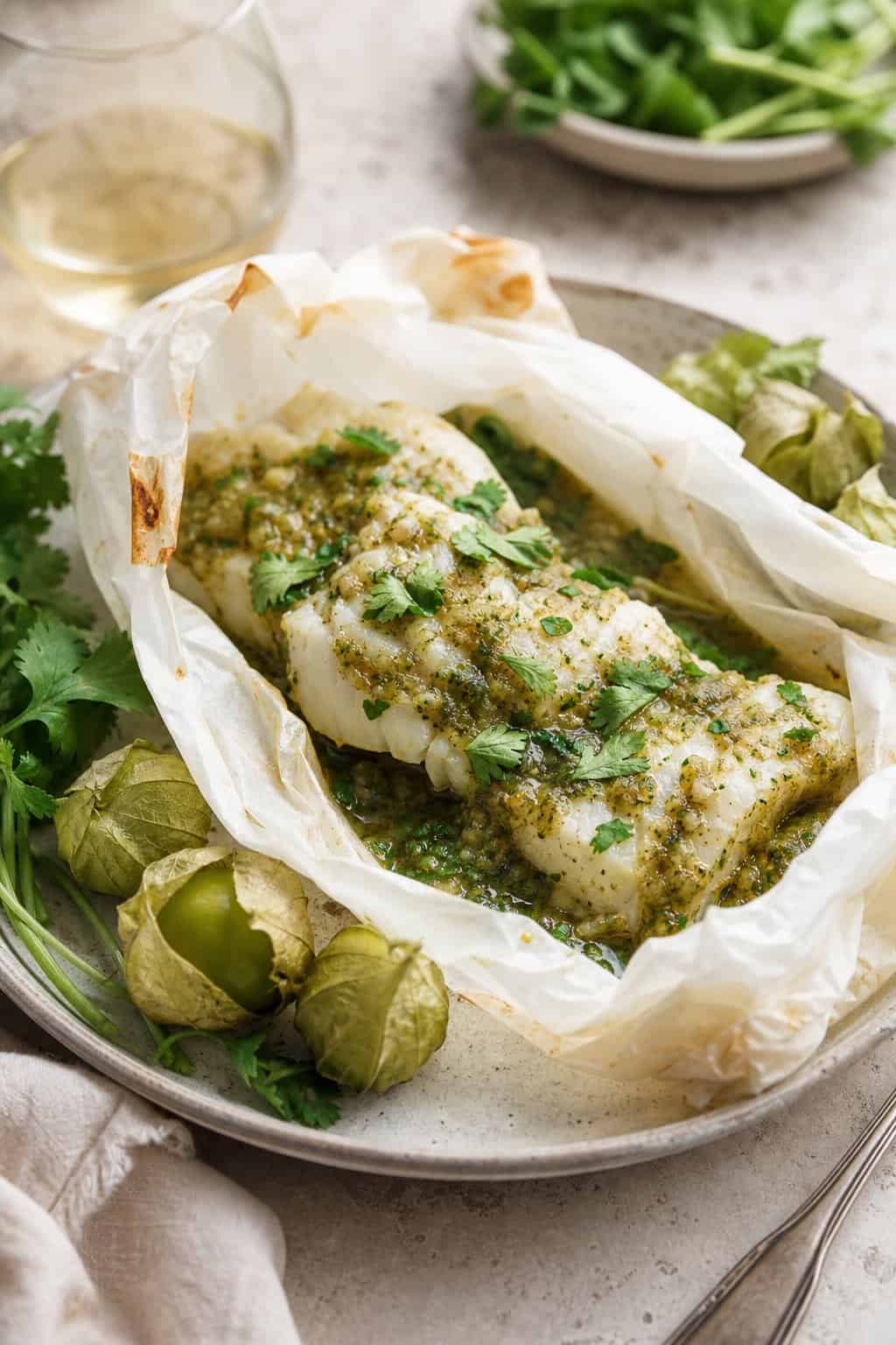 A plated fish dish wrapped in parchment paper with green salsa and garnished with fresh herbs, surrounded by tomatillo husks on a kitchen table.