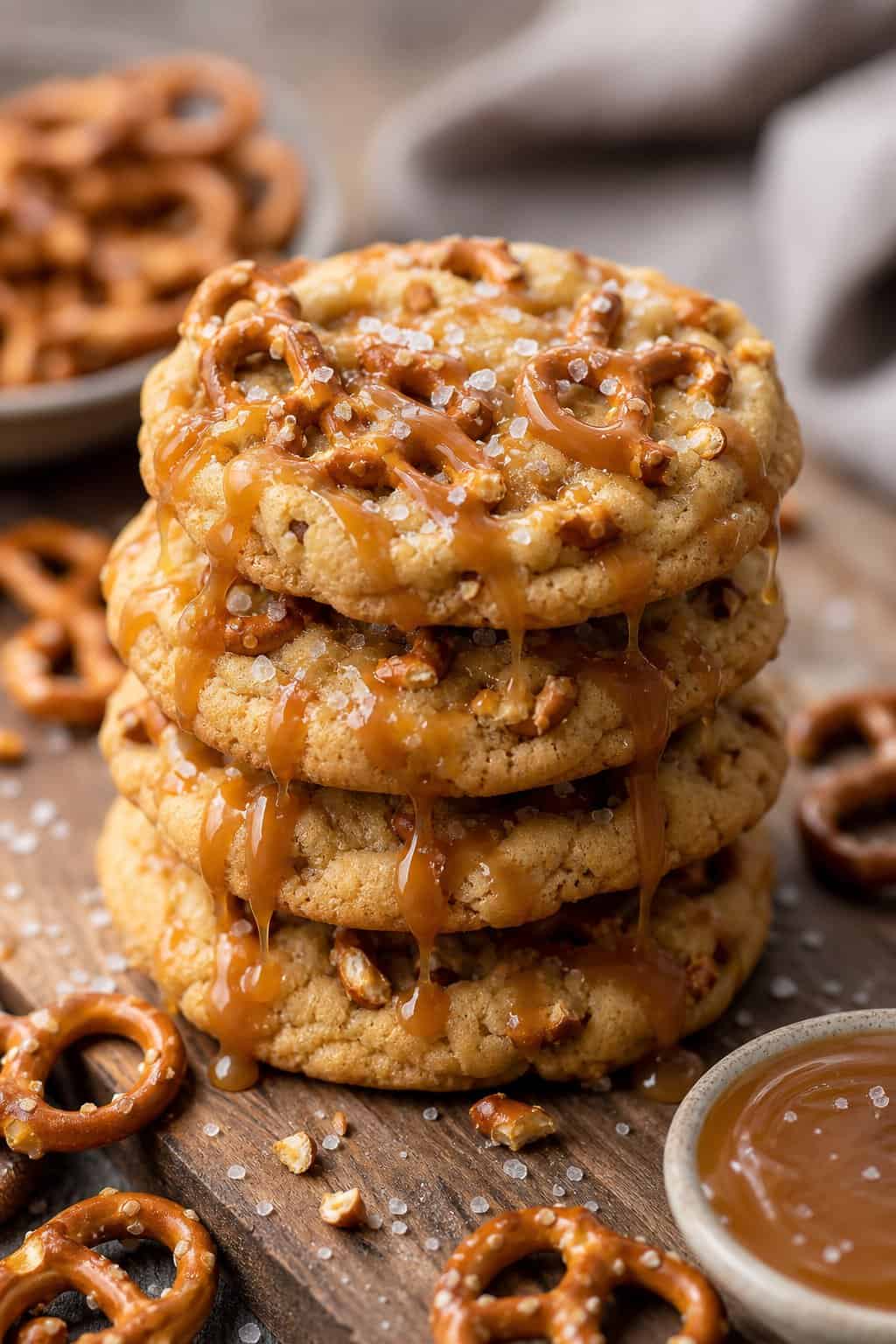 A stack of salted caramel pretzel cookies on a wooden surface with pretzels and caramel sauce nearby.