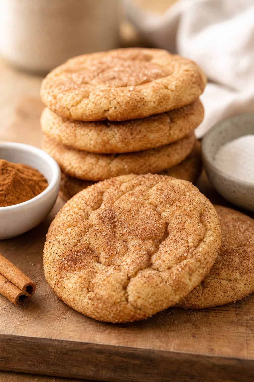 Close-up of freshly baked snickerdoodle cookies coated in cinnamon sugar on a wooden surface with cinnamon and sugar bowls nearby.