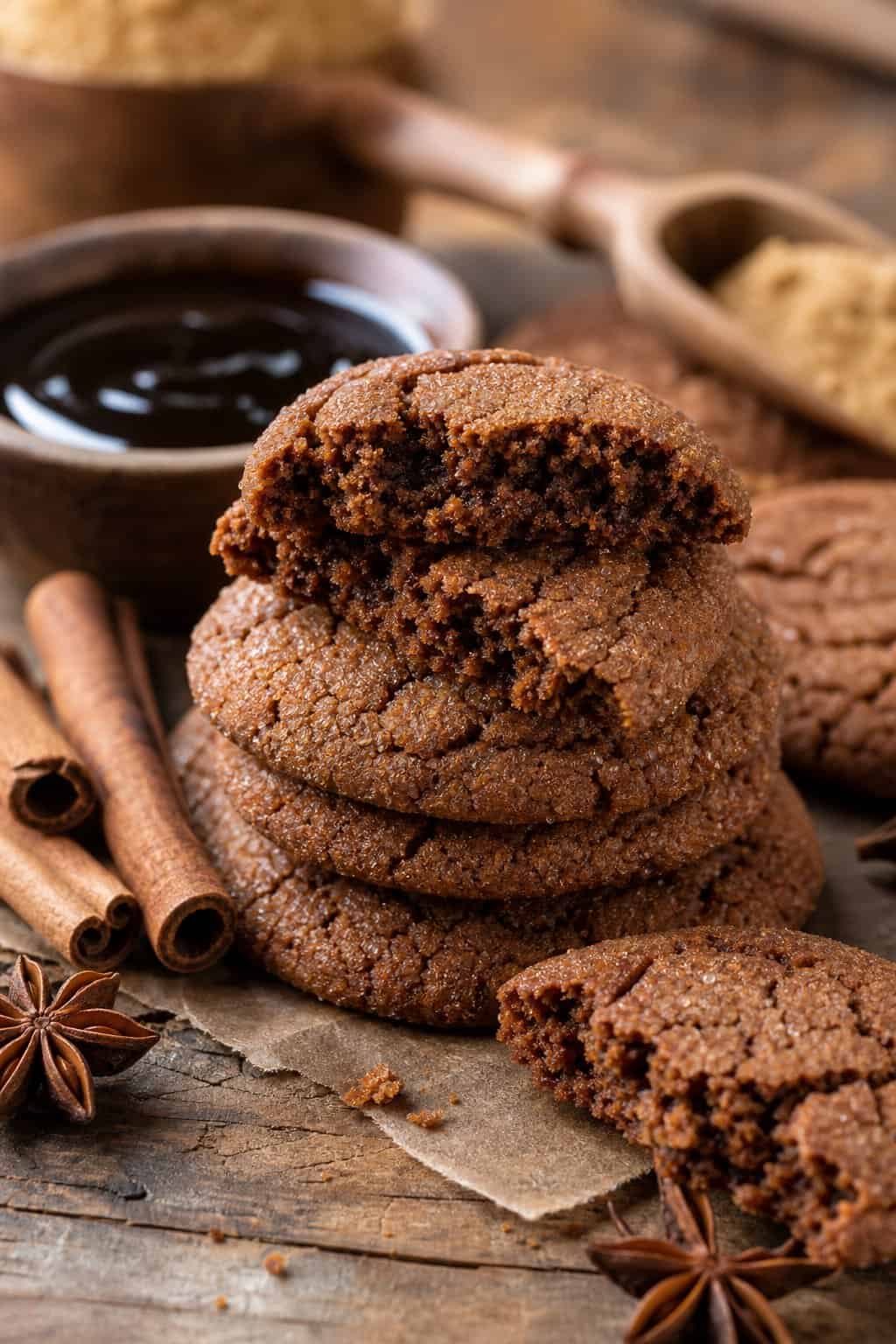 A stack of soft molasses cookies on a wooden surface with cinnamon sticks and molasses nearby.