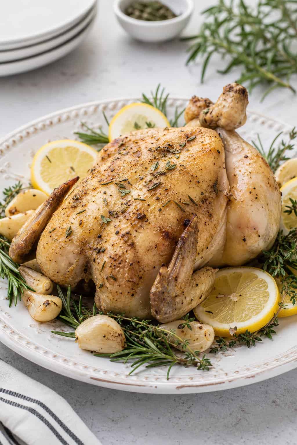 A whole roasted chicken without skin on a white platter garnished with lemon slices, garlic cloves, and fresh herbs.