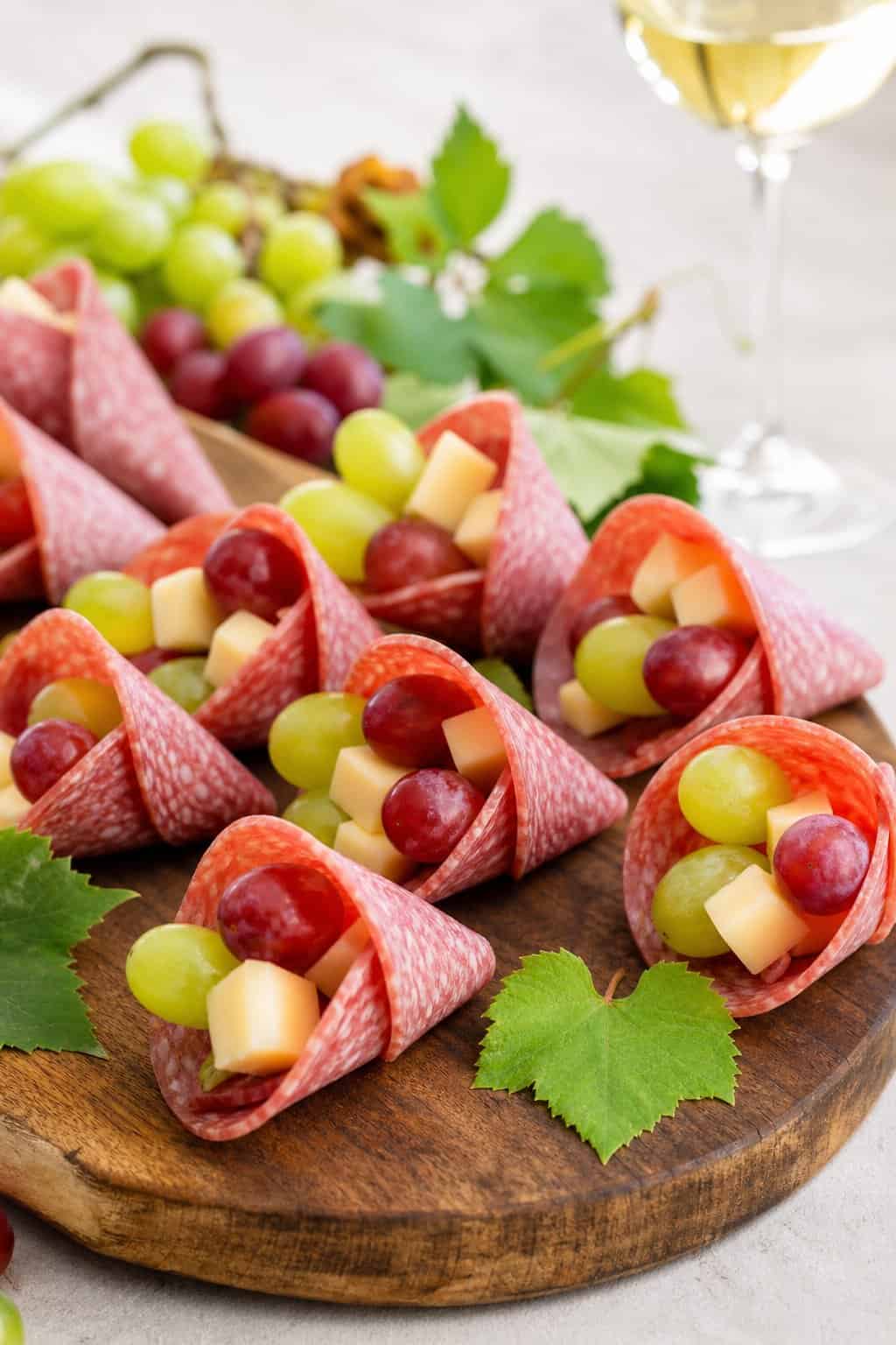 Mini charcuterie cones filled with salami, grapes, and cheese arranged on a wooden serving board.
