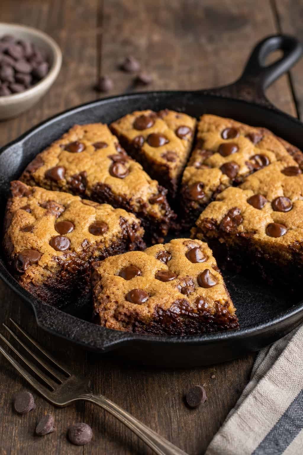 A cast iron skillet filled with a freshly baked brookie, cut into squares, showing a brownie base topped with a chocolate chip cookie layer.
