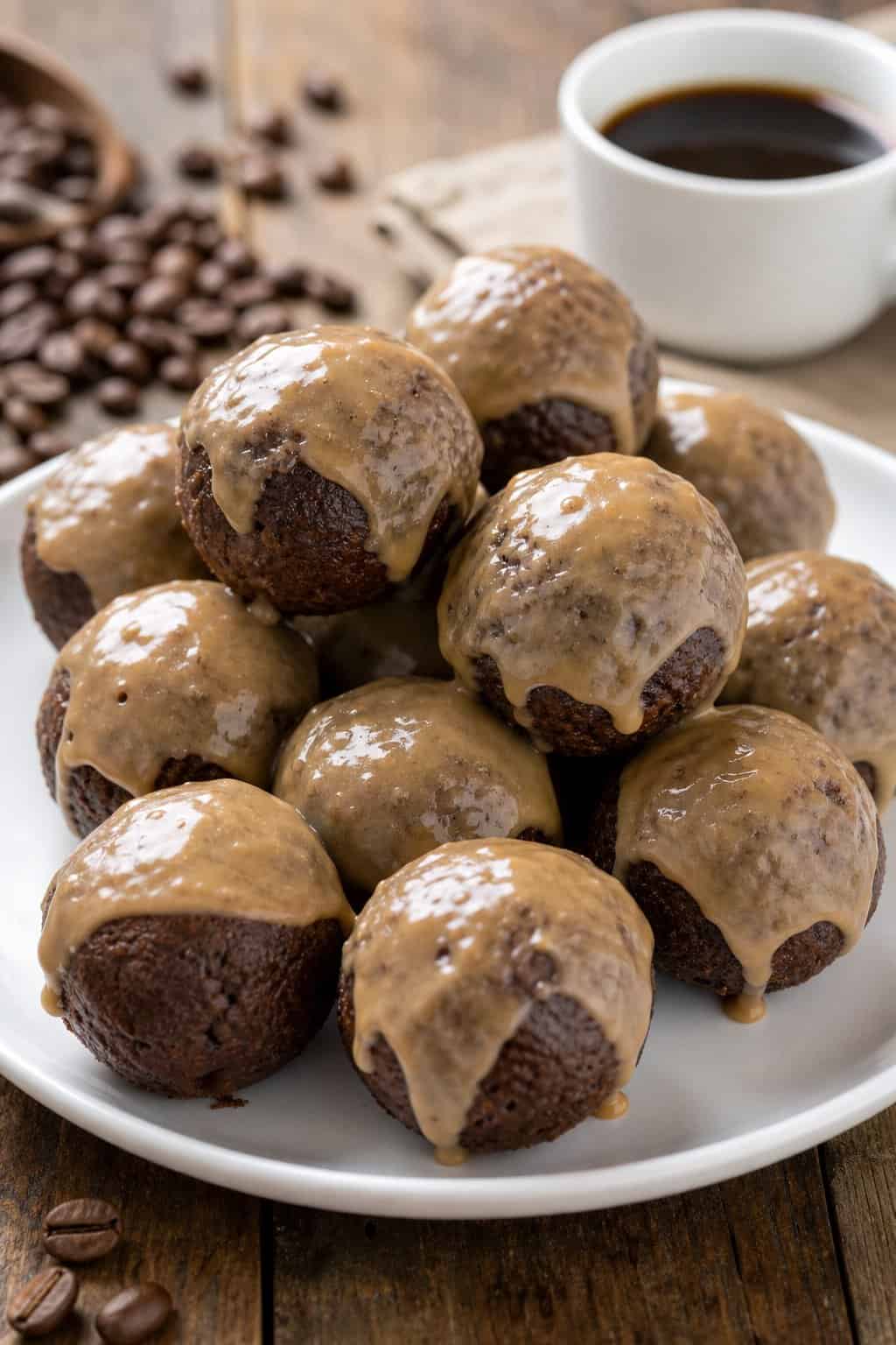 Plate of small brownie donut holes glazed with coffee icing on a wooden table with coffee beans and a cup of coffee in the background.
