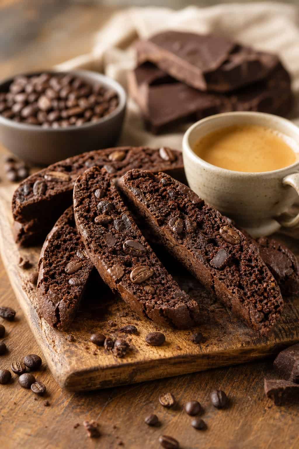 Close-up of espresso brownie biscotti on a wooden board next to a small cup of espresso and scattered coffee beans.