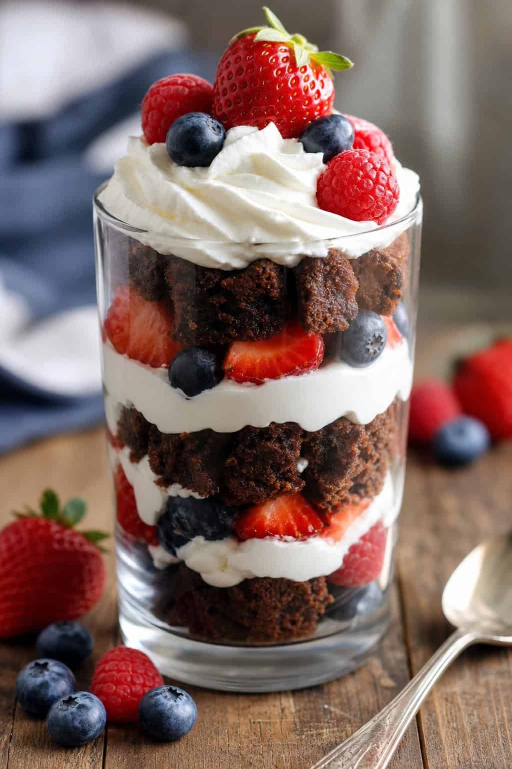 A clear glass parfait layered with brownie pieces, whipped cream, and mixed fresh berries on a wooden table.