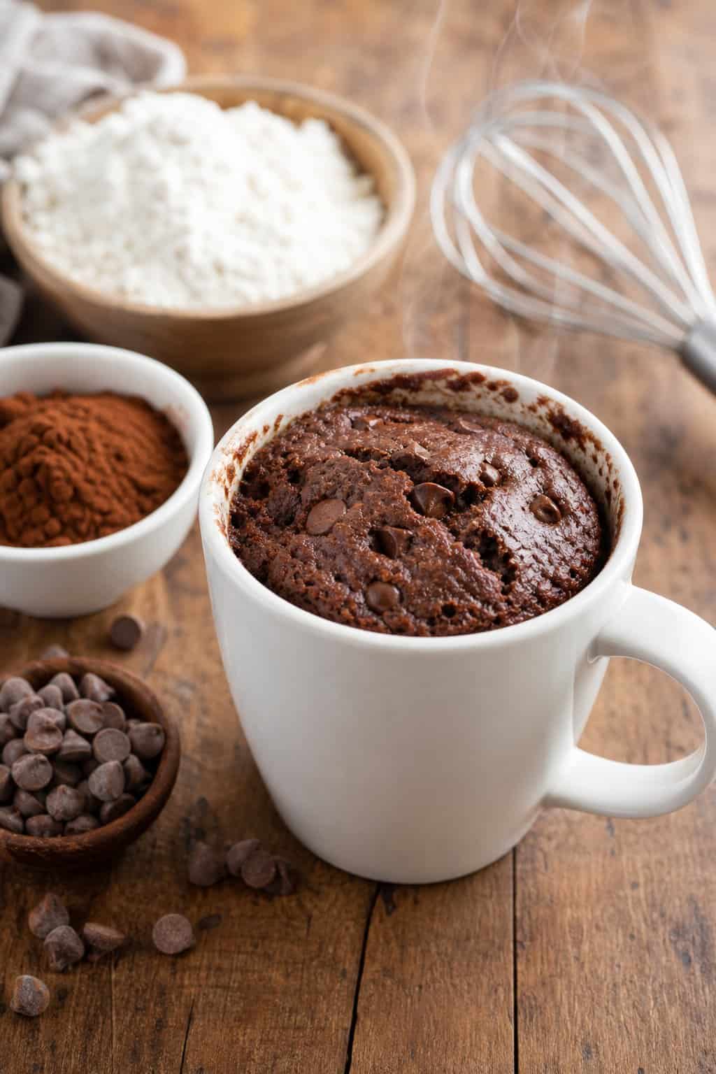 A single chocolate brownie mug cake in a white mug on a wooden table with baking ingredients around it.