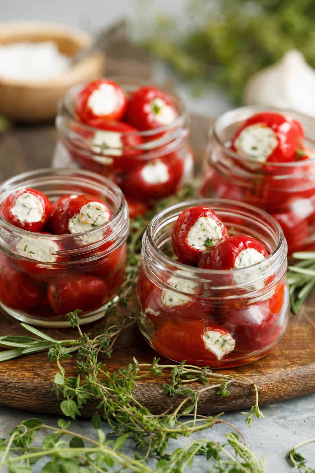 Close-up of glass jars filled with red peppadew peppers stuffed with herbed goat cheese, arranged on a wooden surface with fresh herbs around them.