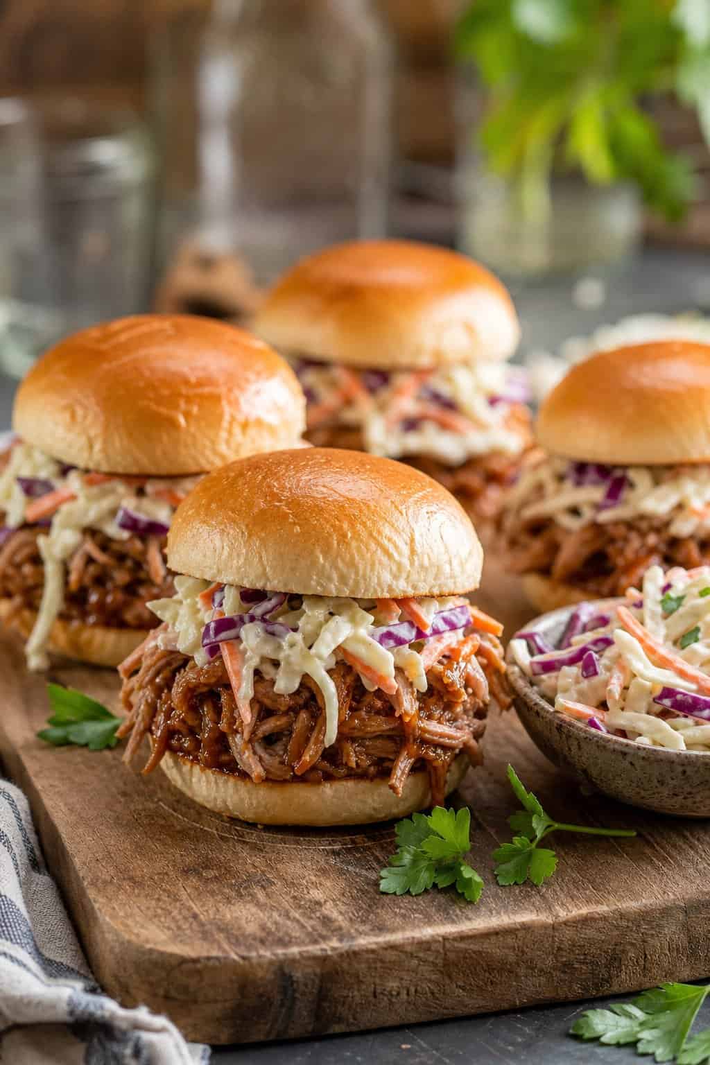 A plate of pulled pork sliders topped with coleslaw on a wooden board in a kitchen setting.