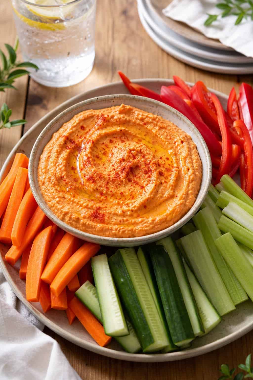 A bowl of roasted red pepper hummus surrounded by fresh vegetable sticks on a wooden table.