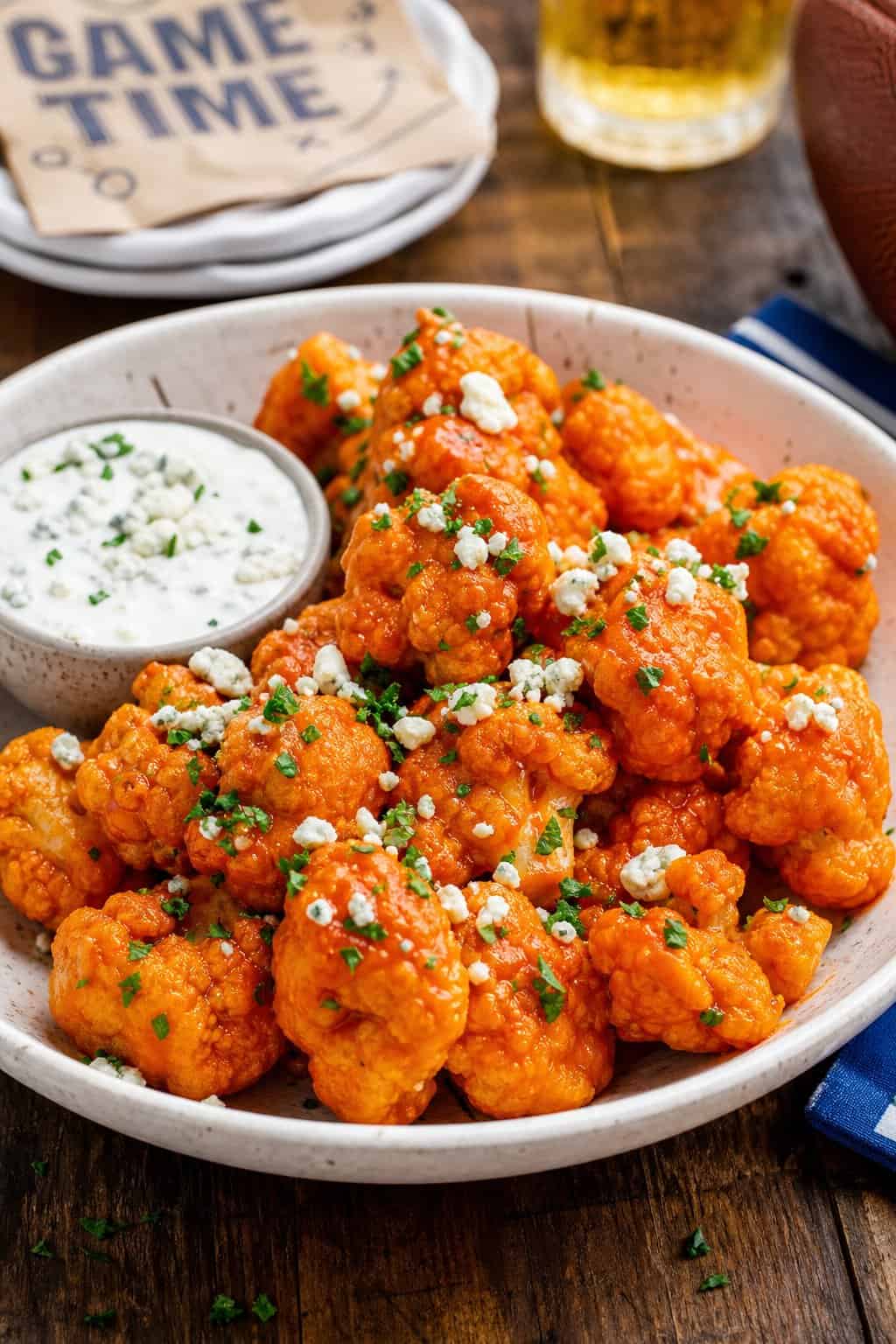 A bowl of buffalo cauliflower bites with blue cheese crumbles and a small cup of blue cheese dressing on a wooden table.