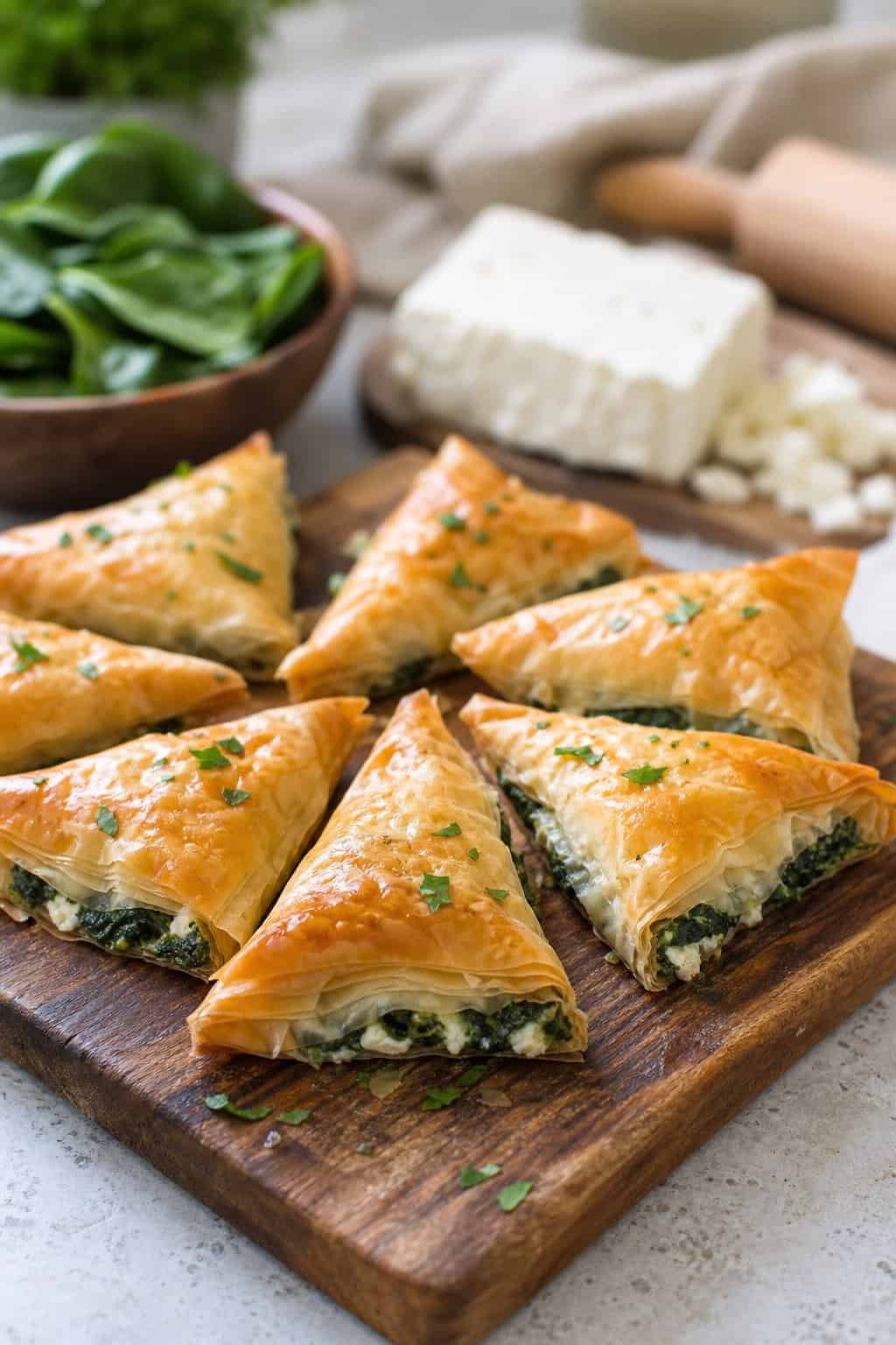 Close-up of golden spinach and feta phyllo triangles arranged on a wooden cutting board with fresh spinach and feta cheese in the background.