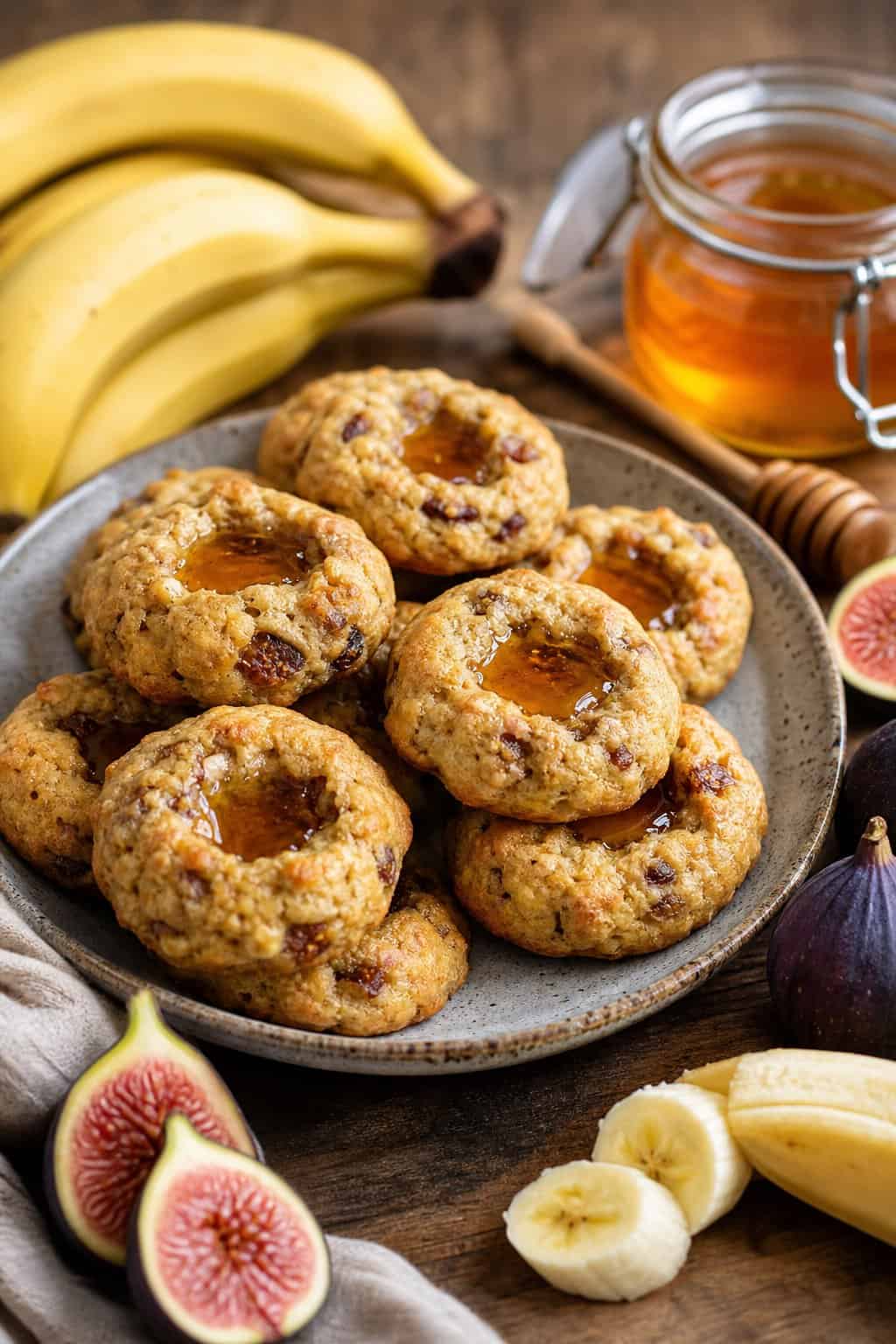 A plate of banana fig and honey thumbprint cookies surrounded by fresh bananas, figs, and a jar of honey on a wooden table.