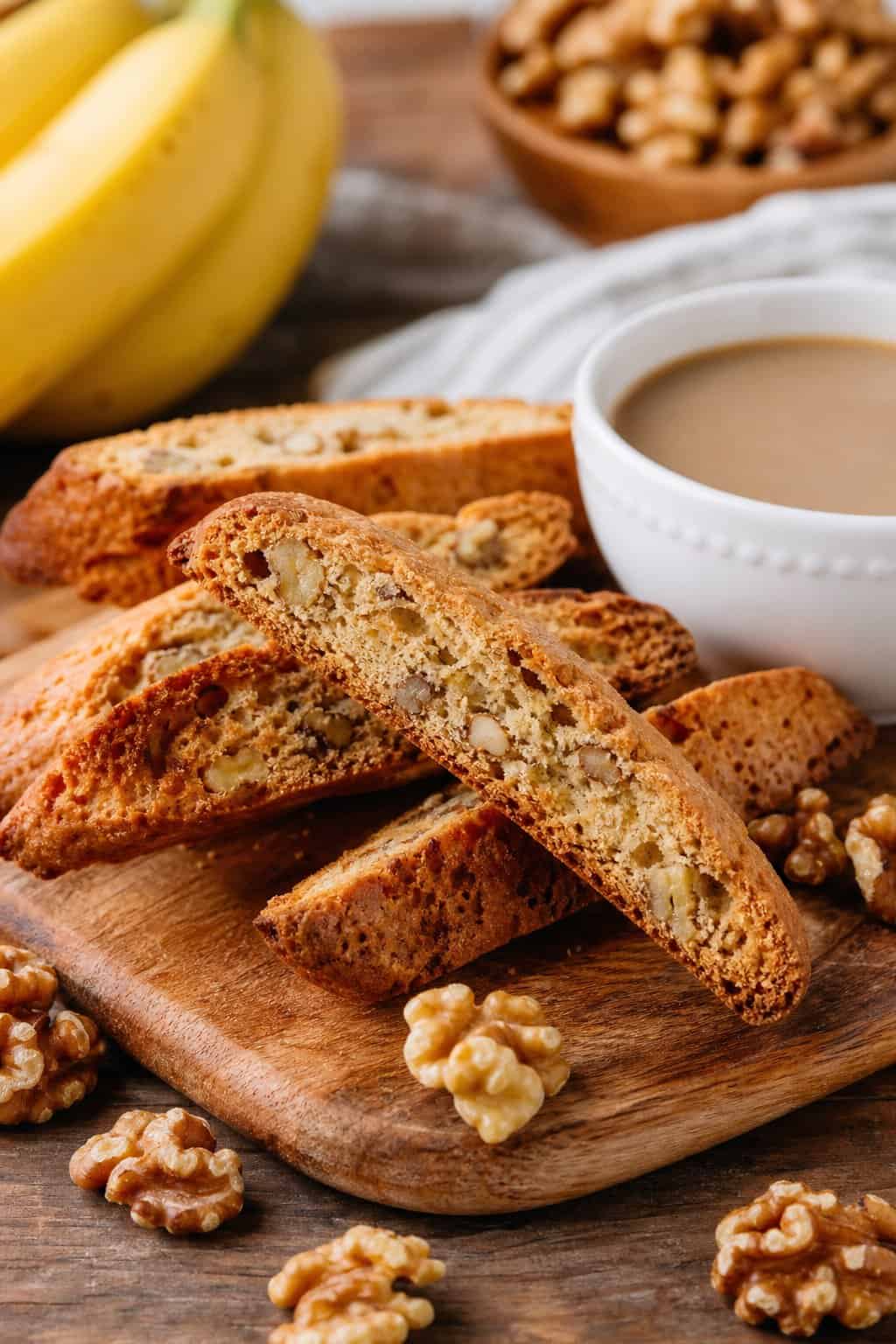 Close-up of banana walnut biscotti on a wooden board with a cup of coffee and bananas in the background.