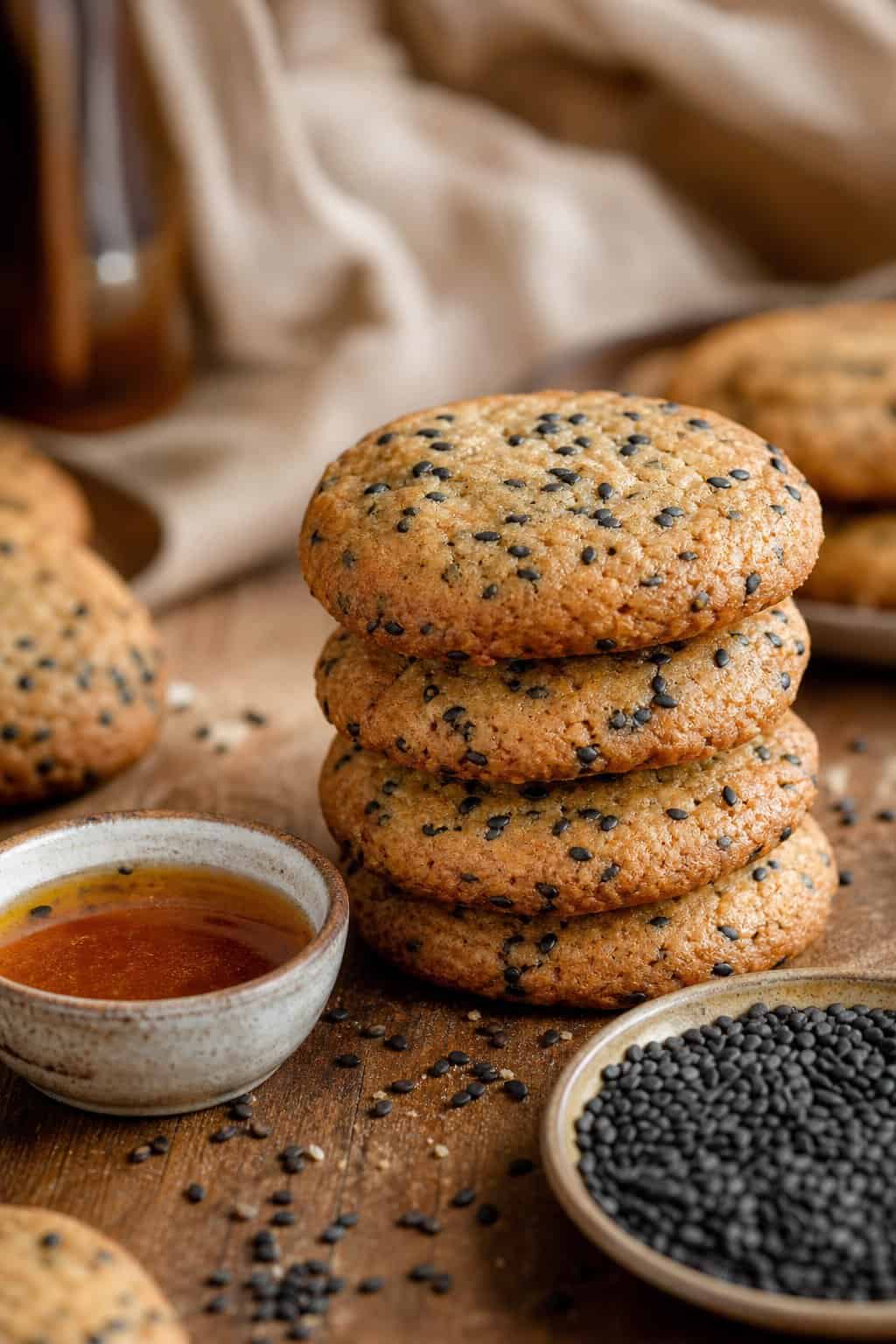 A stack of black sesame brown butter cookies on a wooden surface with scattered sesame seeds and a bowl of melted brown butter nearby.