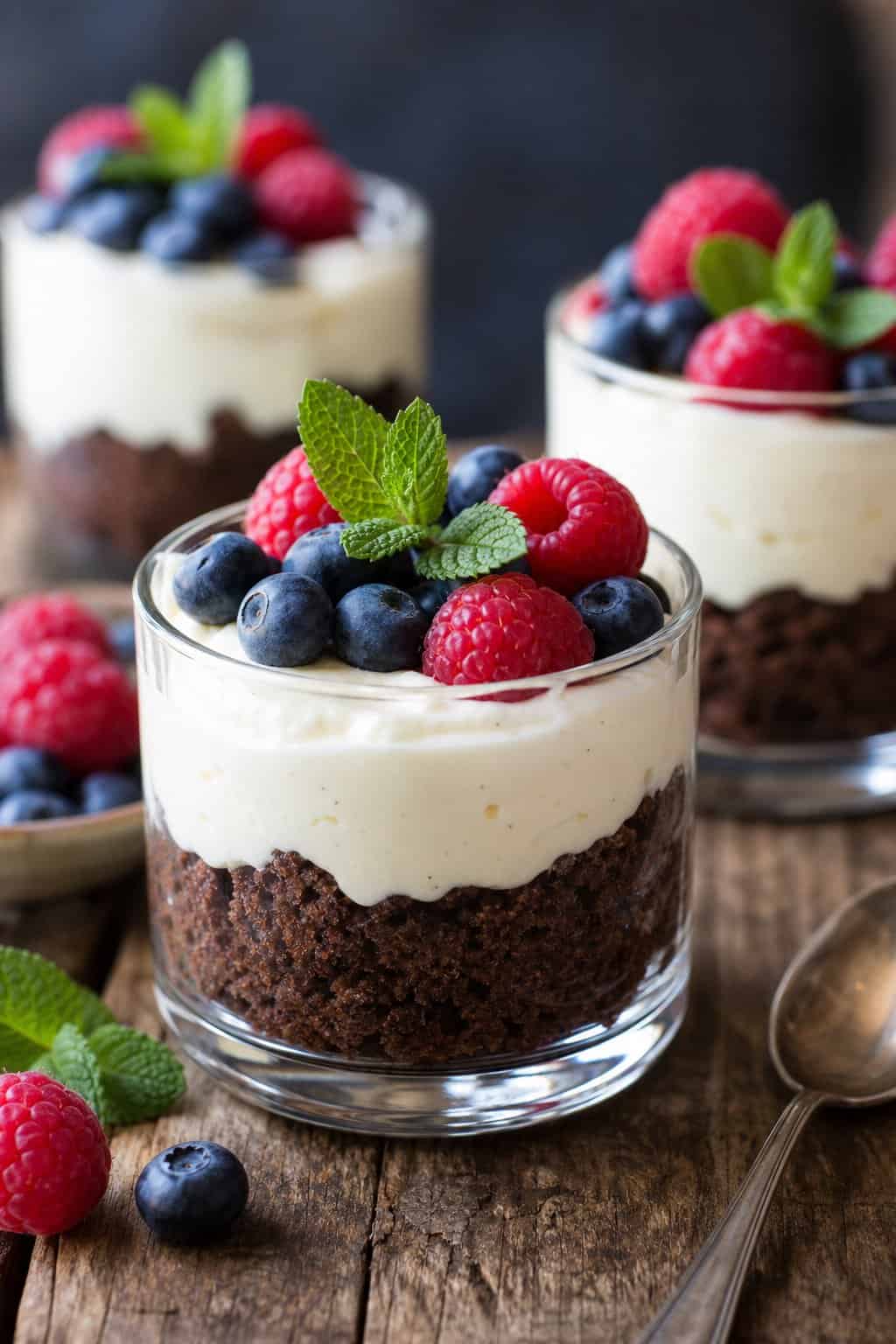 Several glass cups filled with layered brownie crust, cheesecake pudding, and fresh berries on a wooden table.