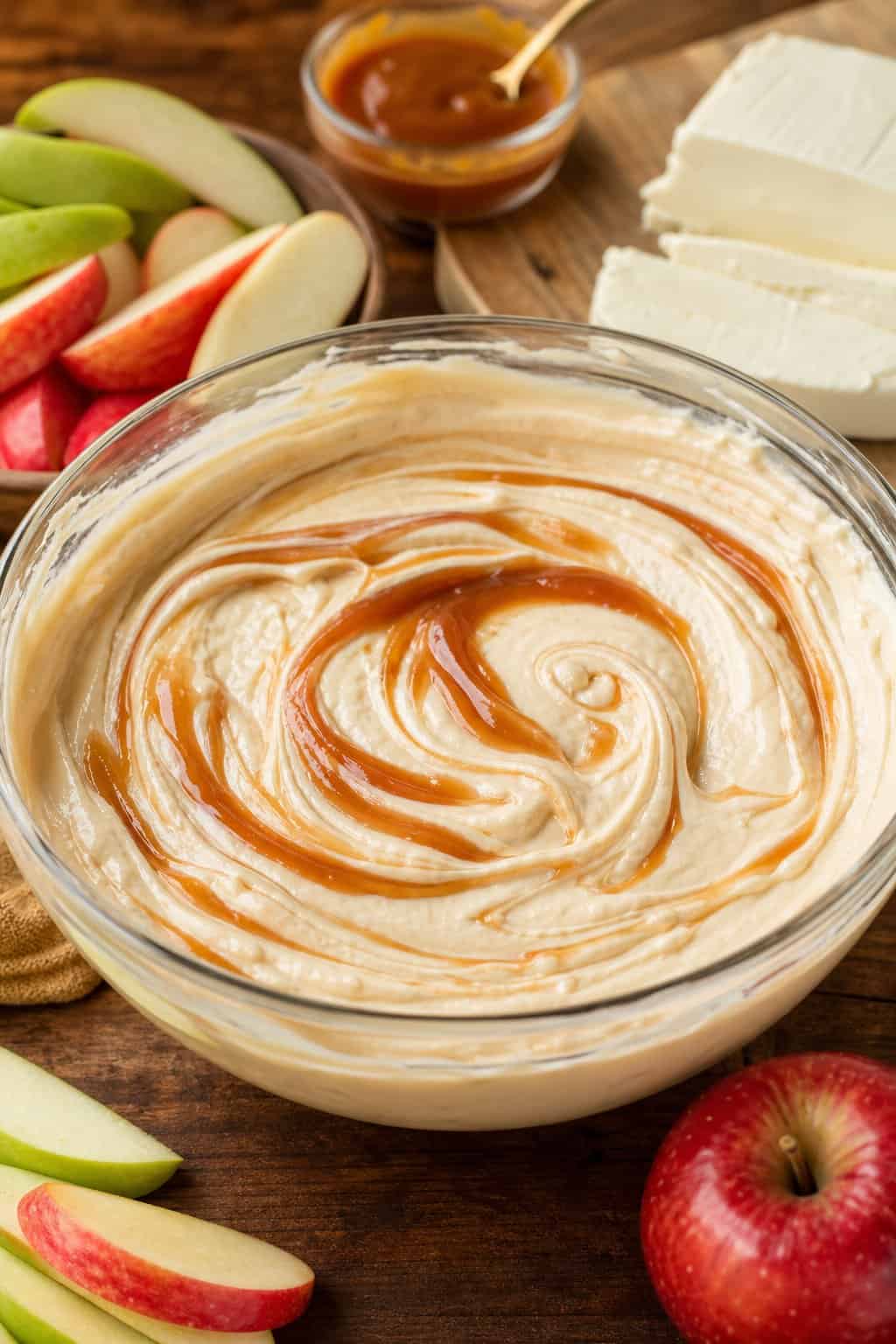 A bowl of creamy caramel apple cheesecake filling surrounded by apple slices, caramel sauce, and cream cheese on a wooden surface.