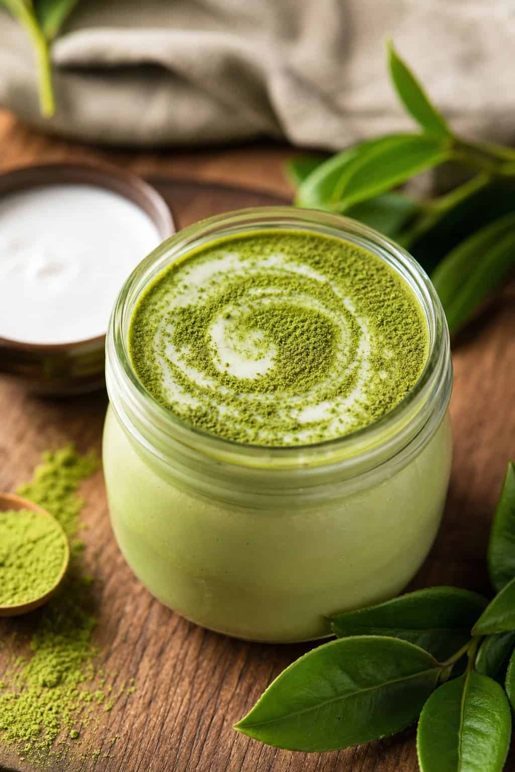 A glass jar filled with creamy matcha cheesecake pudding topped with green matcha powder, placed on a wooden table with matcha leaves and a bowl of coconut milk nearby.