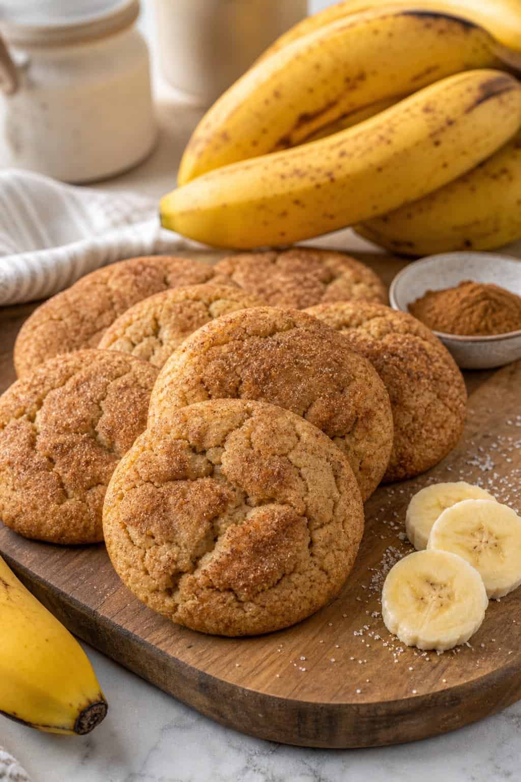 A close-up of freshly baked banana snickerdoodle cookies on a wooden board with ripe bananas and cinnamon nearby.