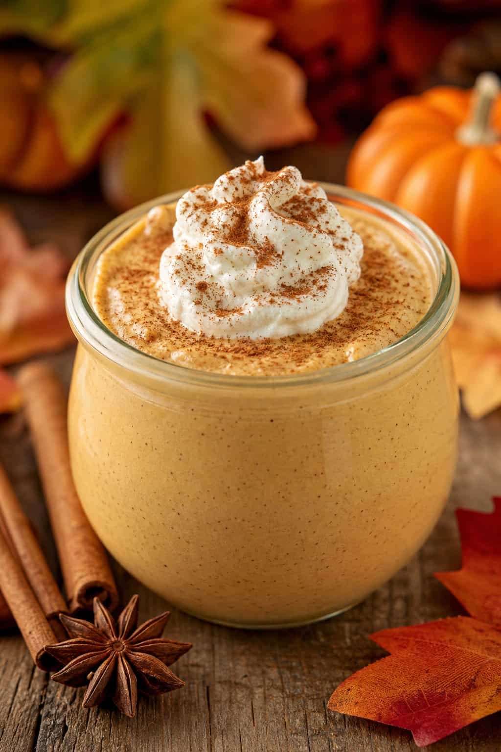 A glass jar filled with pumpkin spice cheesecake pudding topped with whipped cream and cinnamon, surrounded by autumn decorations on a wooden table.
