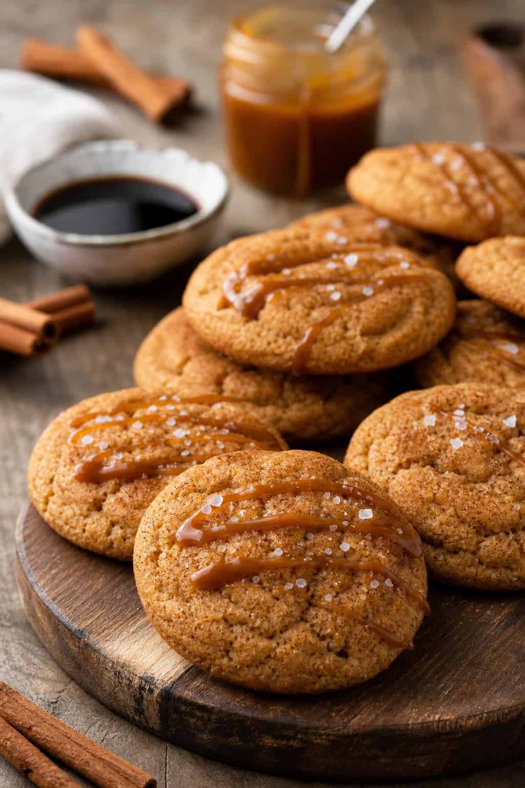 A plate of golden-brown salted caramel soy sauce snickerdoodle cookies with caramel drizzle and sea salt on a wooden surface, surrounded by cinnamon sticks and small bowls of soy sauce and caramel.