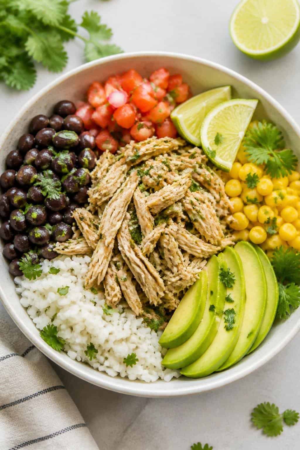 A wide bowl on a wooden table layered with white rice, black beans, corn, shredded cilantro-lime chicken, fanned avocado slices, and a spoonful of bright red pico de gallo.