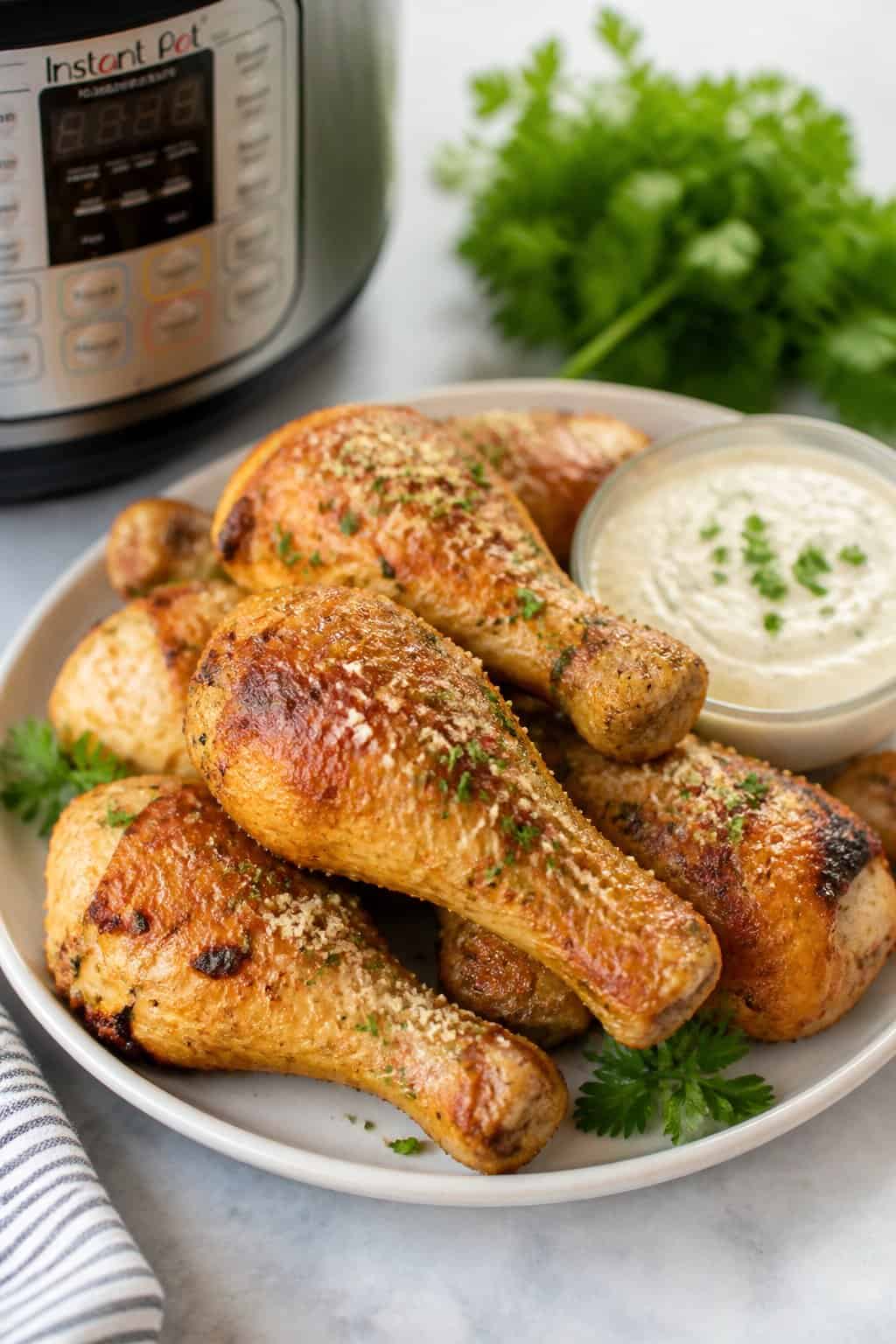 Golden garlic-Parmesan chicken drumsticks on a white plate topped with bright green parsley, with a small white bowl of creamy dipping sauce on a light kitchen table.