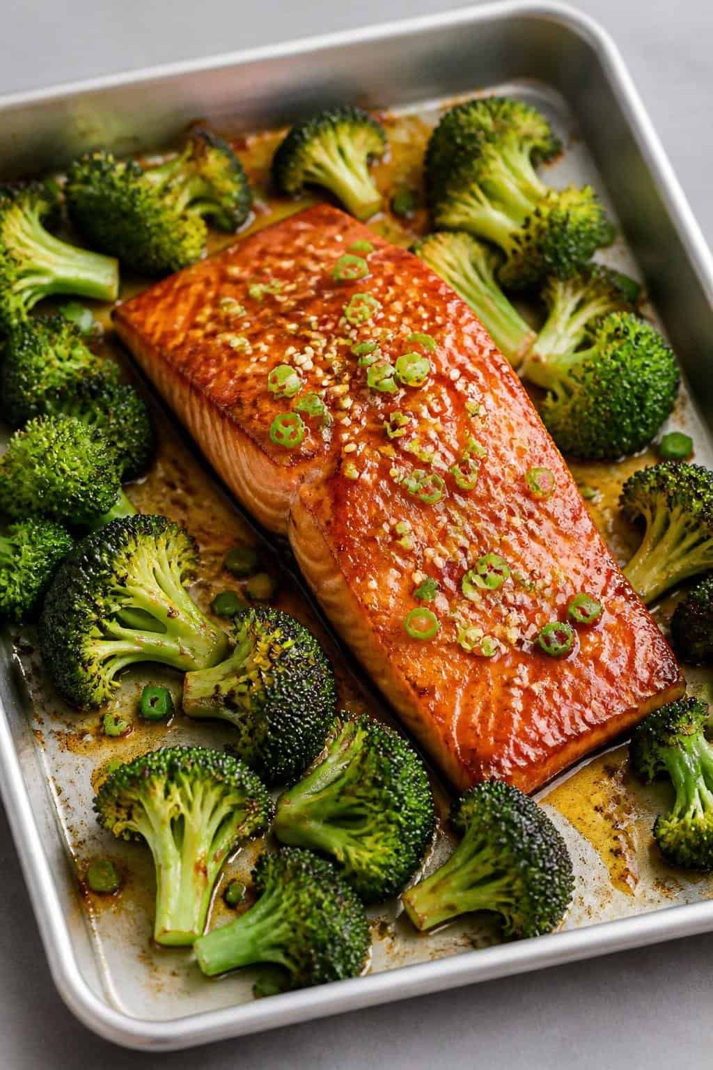 Skin-on salmon fillets glazed in a dark honey-soy sauce resting beside deeply charred broccoli florets on a foil-lined sheet pan.