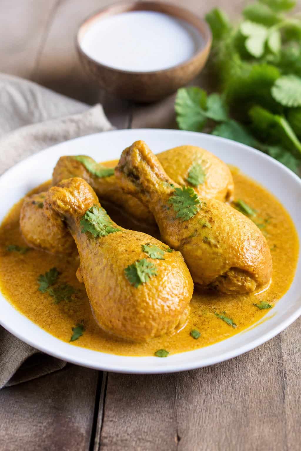 Lightly bronzed chicken drumsticks in a pale golden coconut curry sauce in a white baking dish, topped with fresh cilantro leaves, on a wooden surface.
