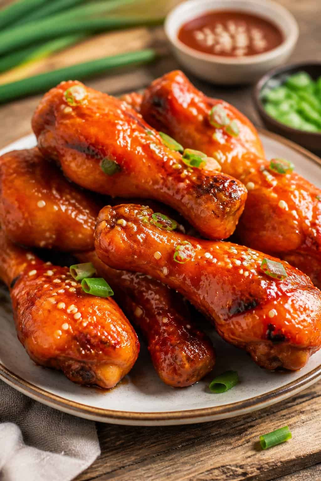 Shiny orange-glazed chicken drumsticks on a white round plate, topped with thin green onion rounds, on a warm wooden table.