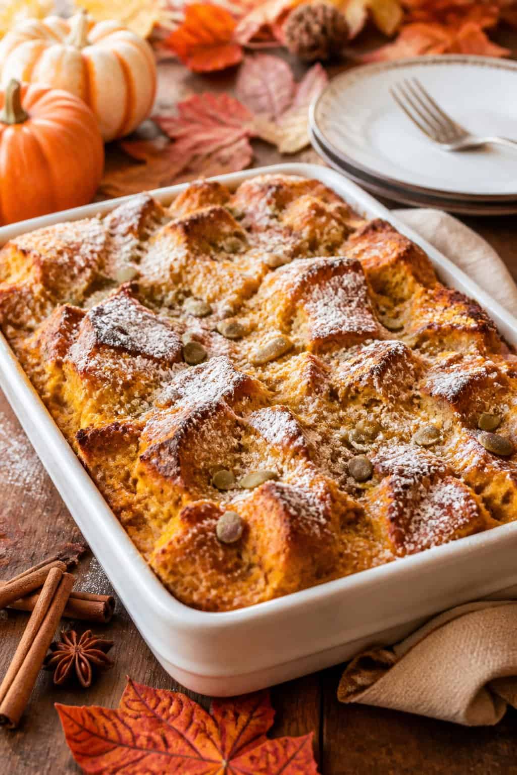 A pumpkin-spice breakfast casserole in a deep ceramic dish on a wooden table with small decorative pumpkins, cinnamon sticks, and rust-colored autumn leaves in the background.