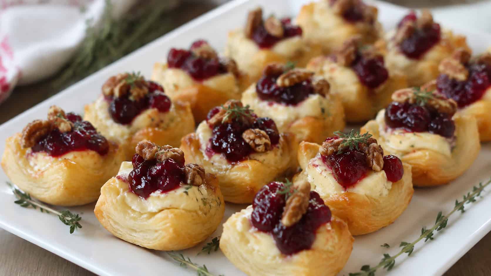 golden cranberry brie bites arranged on a white platter in a wide horizontal shot, ruby cranberry and toasted pecans on top, sprigs of thyme nearby, soft natural light, shallow depth of field, landscape orientation