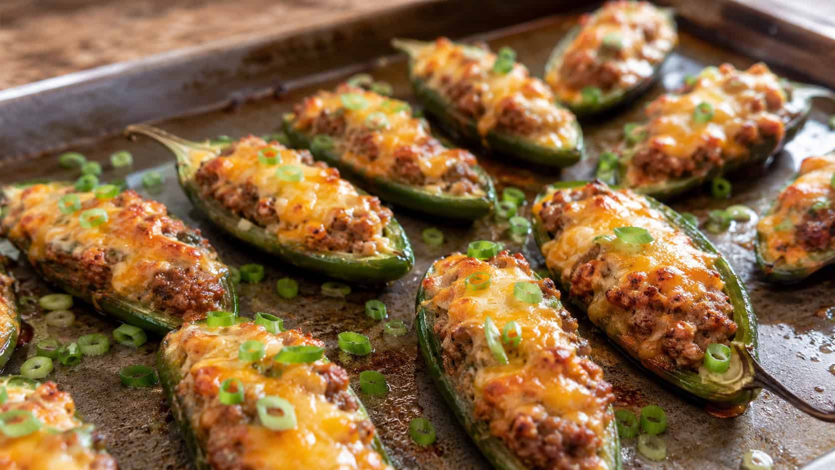 sausage stuffed jalapeno halves on a baking sheet in a wide horizontal shot, melted cheese bubbling and golden, scattered green onion, warm natural light, shallow depth of field, landscape orientation