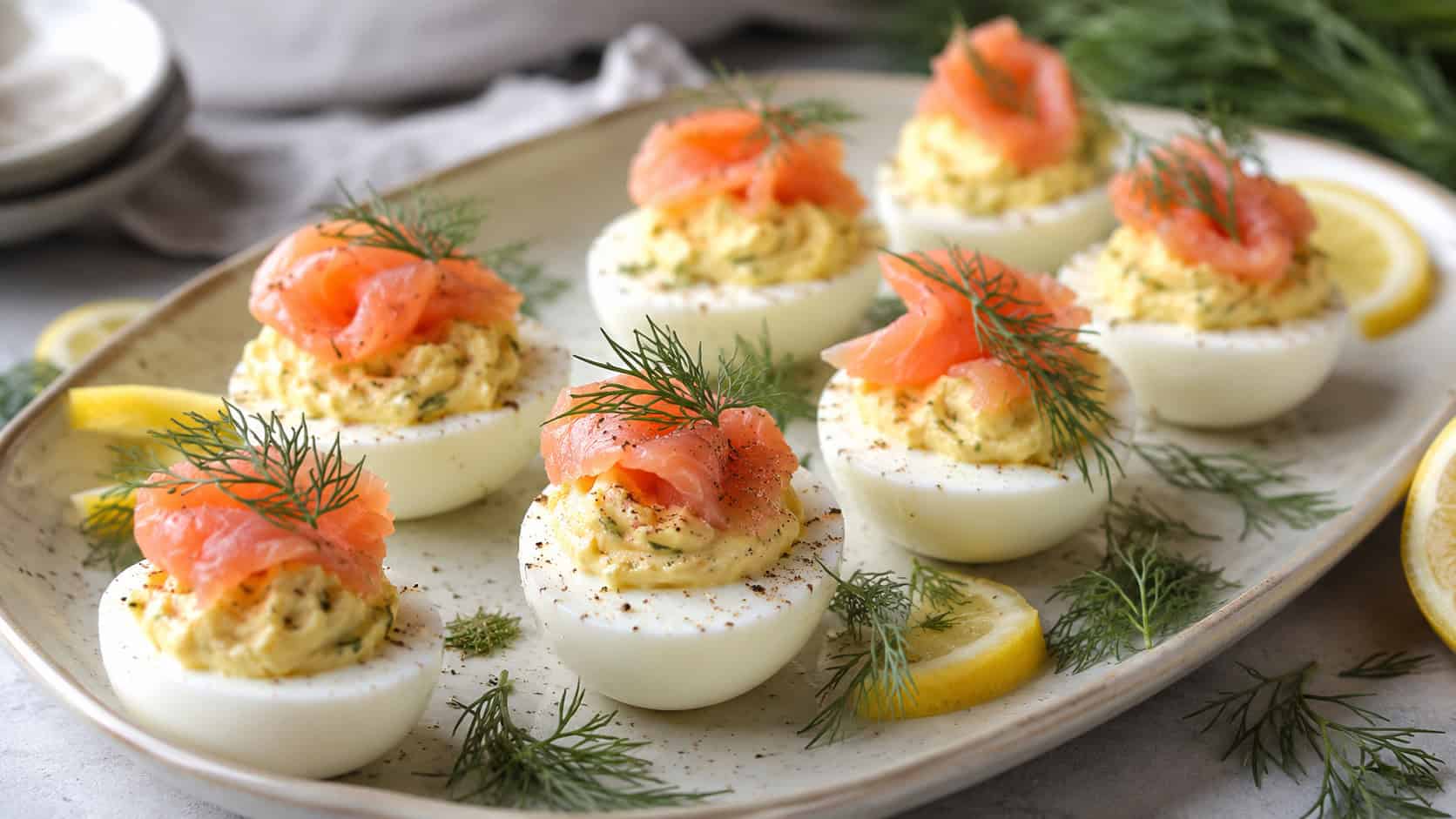 deviled eggs topped with smoked salmon and dill on a pale ceramic platter in a wide horizontal shot, lemon and dill sprigs scattered nearby, soft natural light, shallow depth of field, landscape orientation