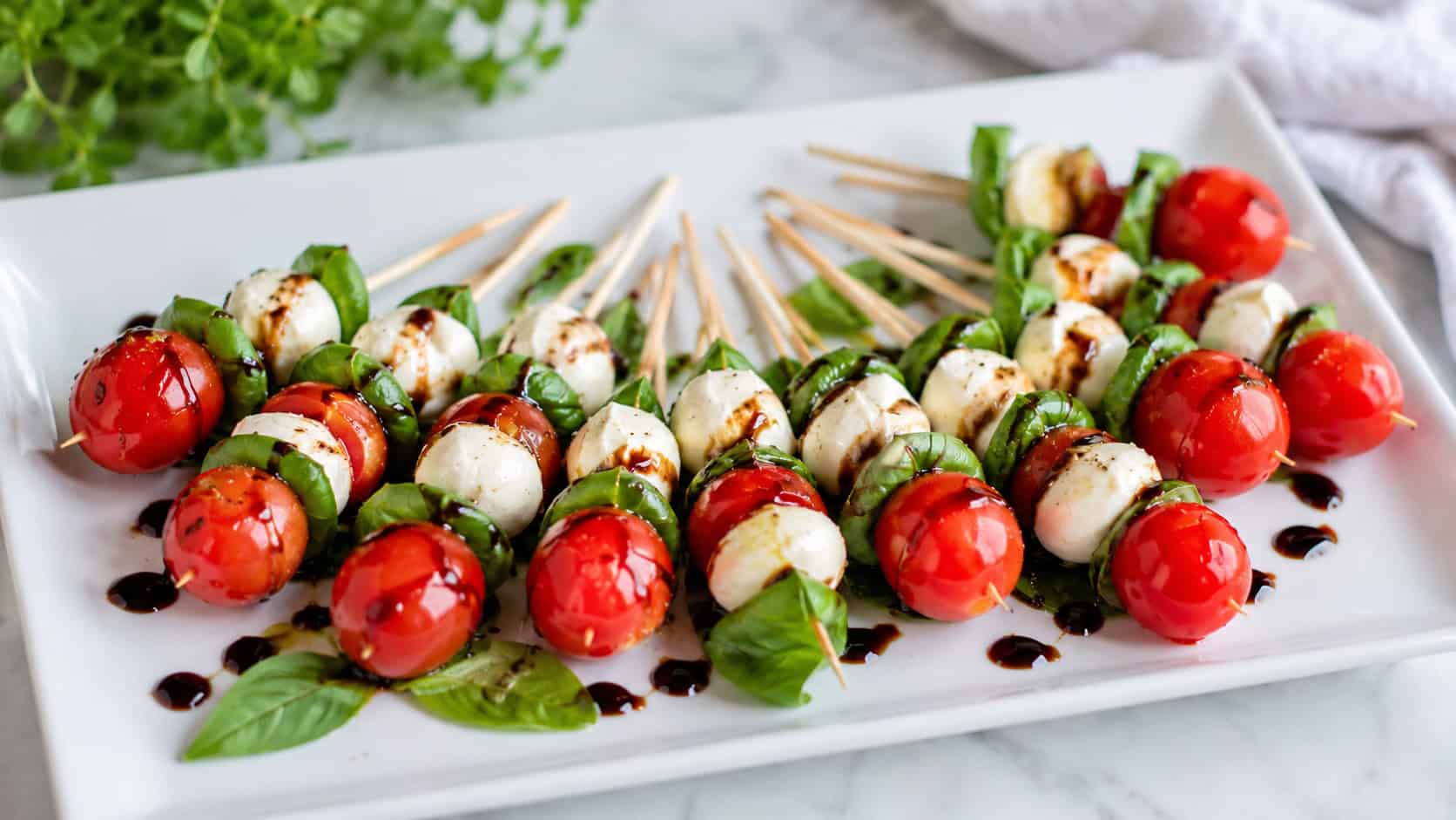 mini caprese skewers fanned across a white rectangular platter in a wide horizontal shot, glossy balsamic drizzle, fresh basil scattered around, bright natural light, shallow depth of field, landscape orientation