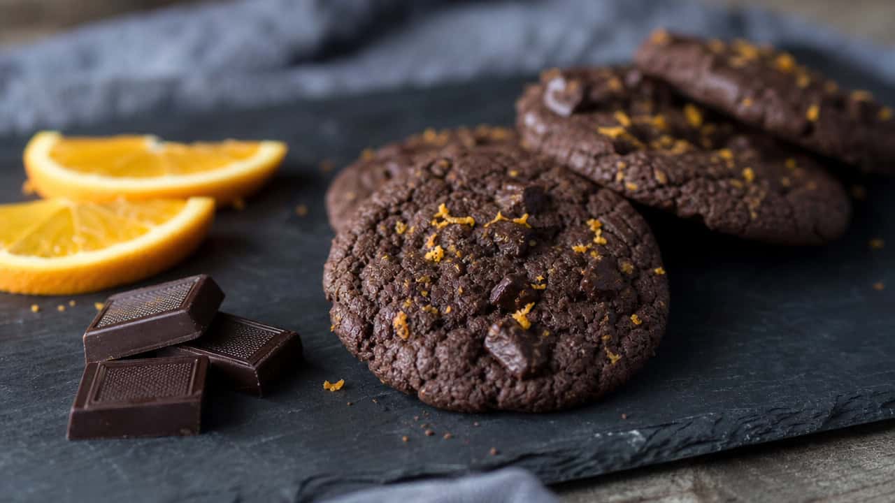 dark chocolate cookies flecked with orange zest on a slate board in a wide horizontal shot, orange slices and a few chocolate squares beside them, moody natural light, shallow depth of field, landscape orientation