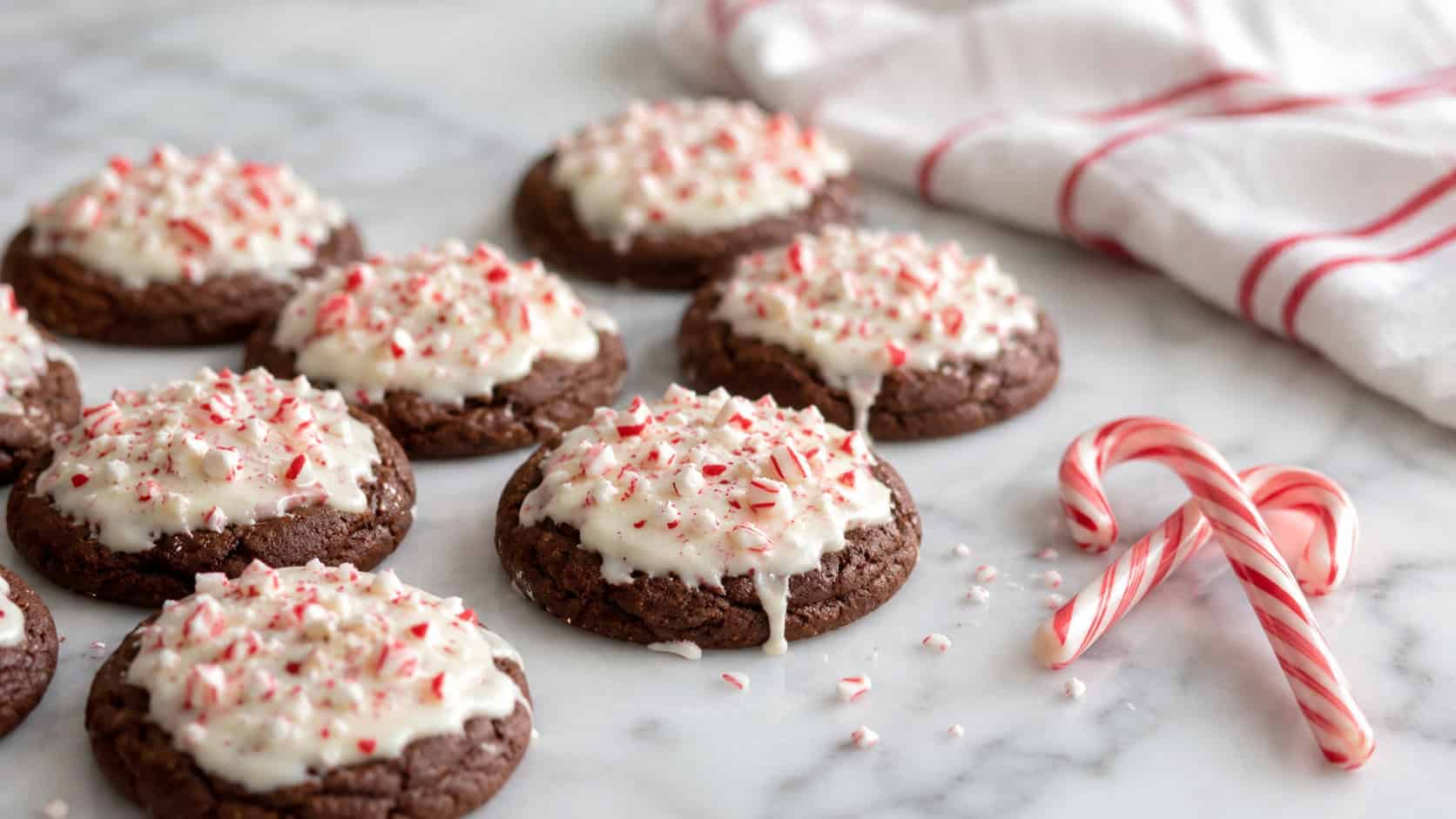 chocolate cookies topped with white chocolate and crushed red candy cane pieces on a white marble counter in a wide horizontal shot, a striped tea towel and full candy canes beside them, soft natural light, shallow depth of field, landscape orientation
