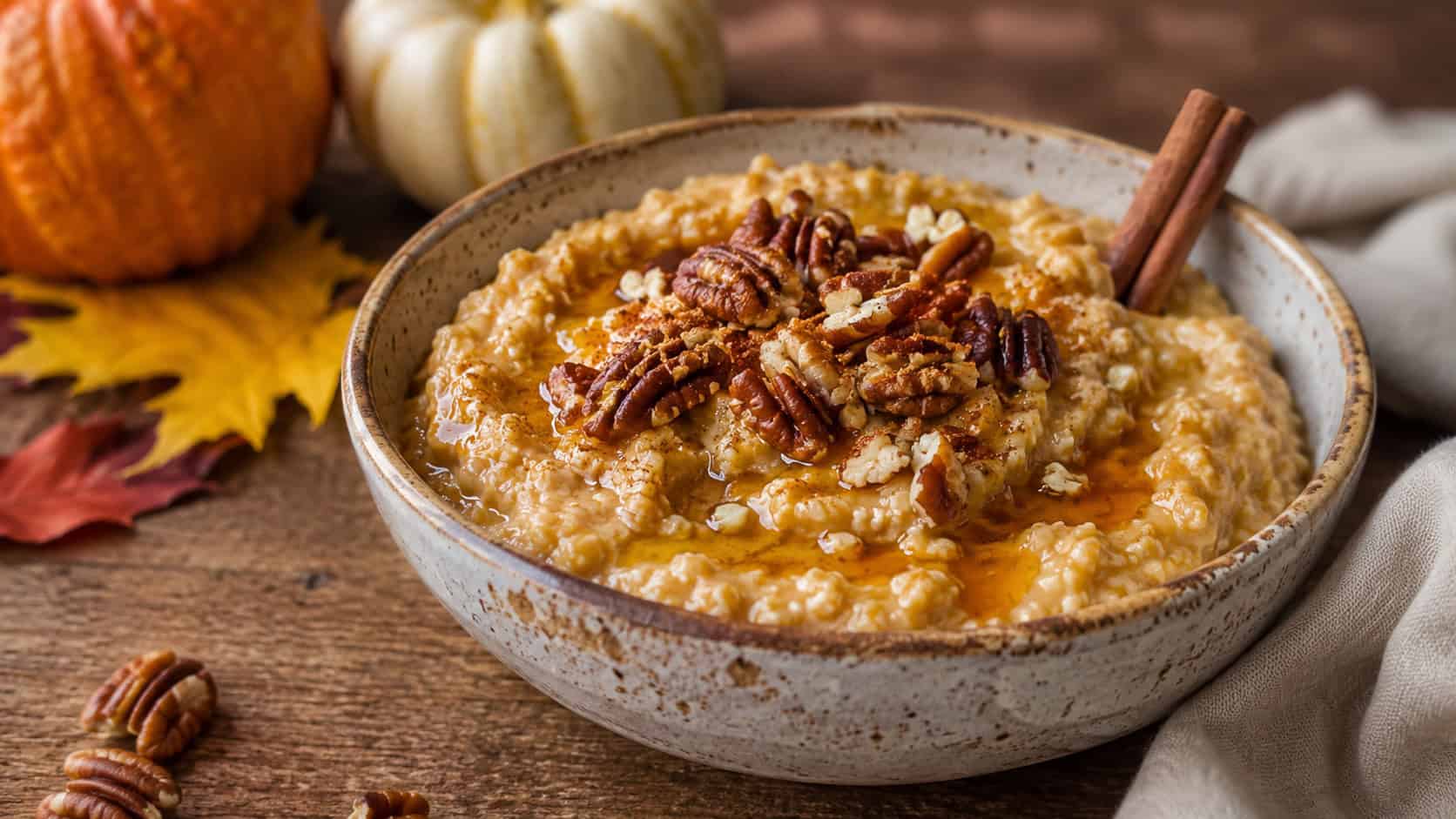 a creamy bowl of pumpkin spice oatmeal topped with toasted pecans and a maple drizzle in a wide horizontal shot, served in a rustic bowl with a cinnamon stick, natural light, shallow depth of field, landscape orientation