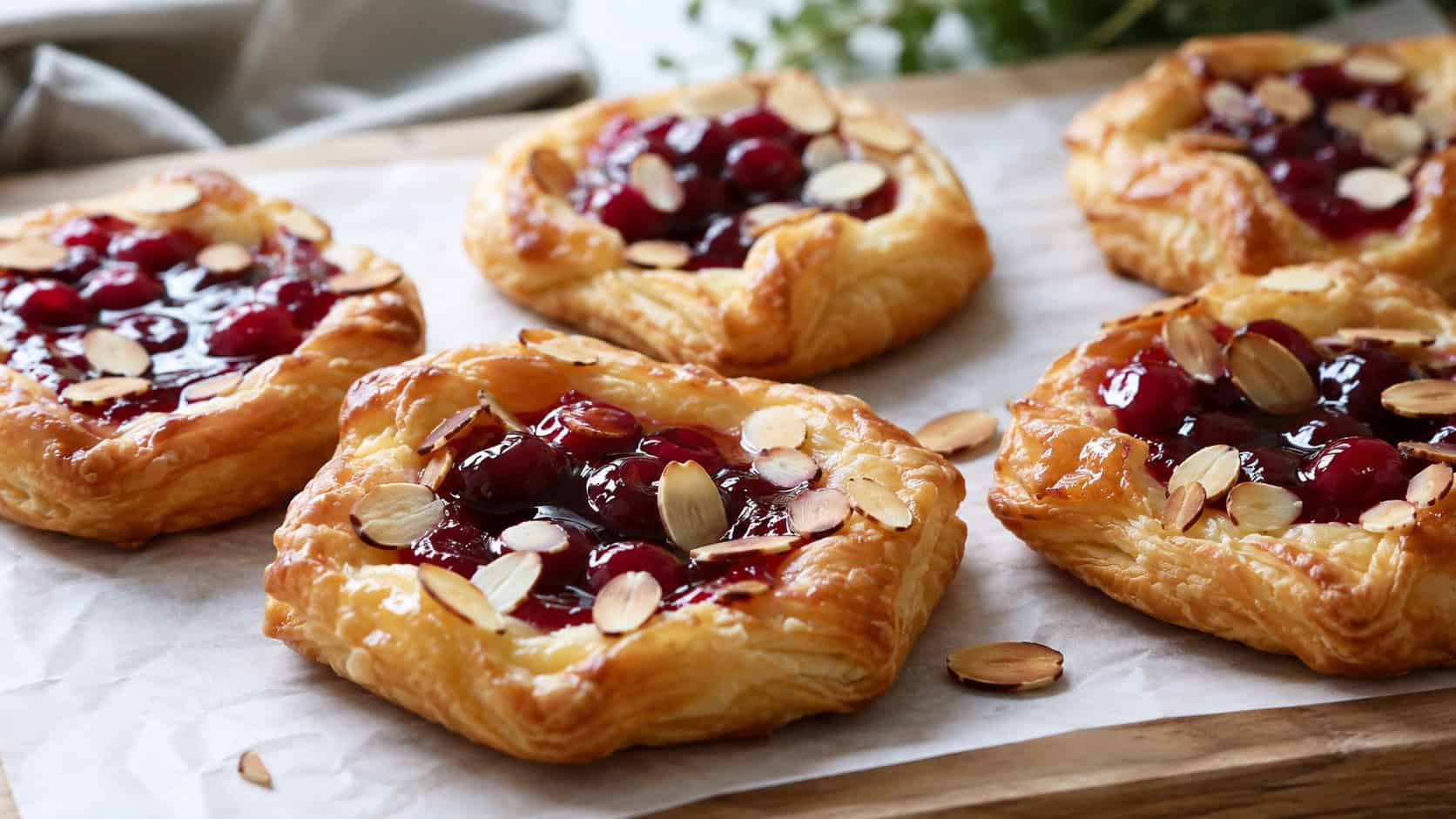 cherry almond danishes with golden puff pastry, glossy cherry centers, and toasted almonds in a wide horizontal shot, arranged on a parchment-lined board, natural light, shallow depth of field, landscape orientation
