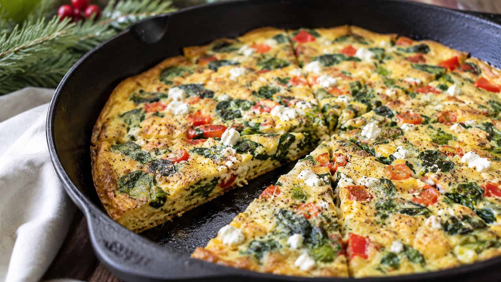 a holiday frittata flecked with red peppers, green spinach, and white feta in a wide horizontal shot, sliced into wedges in a cast iron skillet, natural light, shallow depth of field, landscape orientation