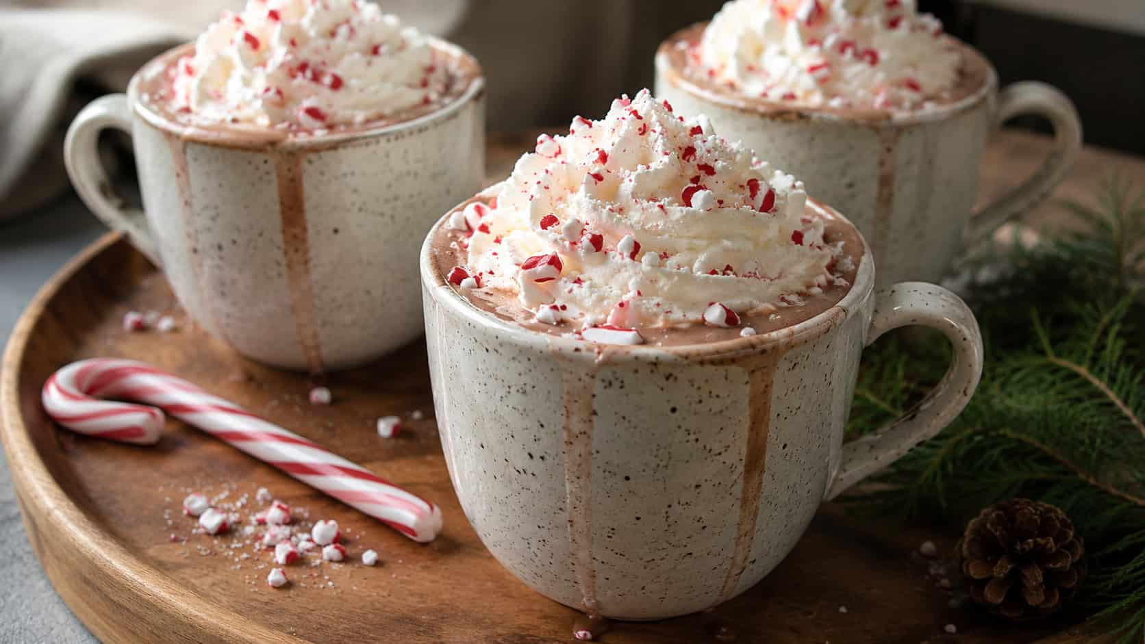 mugs of peppermint hot chocolate topped with whipped cream and crushed candy canes in a wide horizontal shot, arranged on a wooden tray with a candy cane, natural light, shallow depth of field, landscape orientation