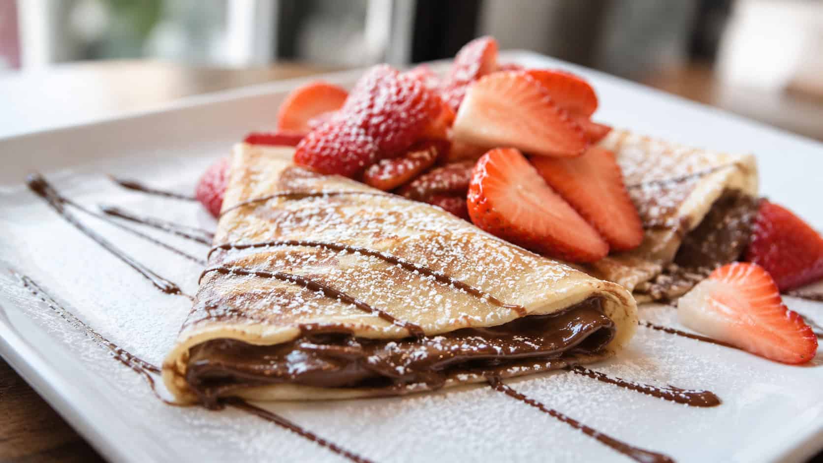 folded Nutella crepes topped with fresh strawberries and a dusting of powdered sugar in a wide horizontal shot, plated on white with a chocolate drizzle, natural light, shallow depth of field, landscape orientation