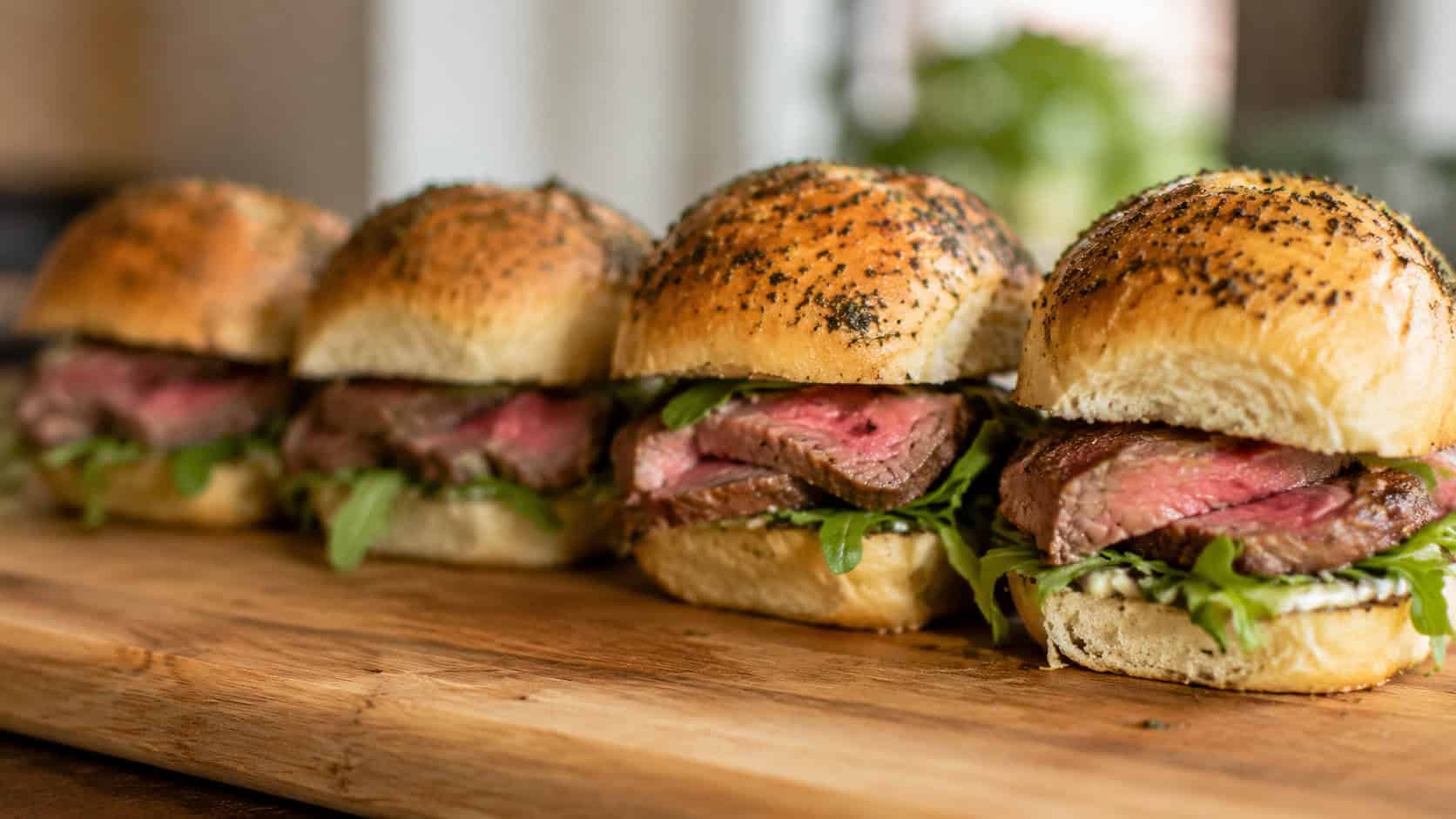 herb crusted prime rib sliders lined up on a wooden board in a wide horizontal shot, pink sliced beef and fresh greens peeking out, soft natural light, shallow depth of field, landscape orientation