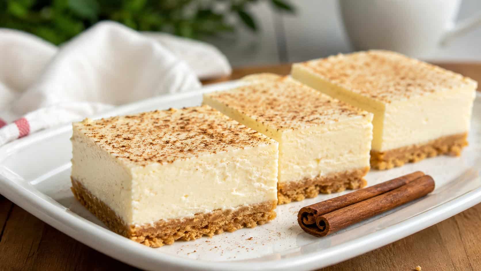 creamy eggnog cheesecake bars dusted with nutmeg on a white plate in a wide horizontal shot, golden crust visible, cinnamon sticks beside them, soft natural light, shallow depth of field, landscape orientation