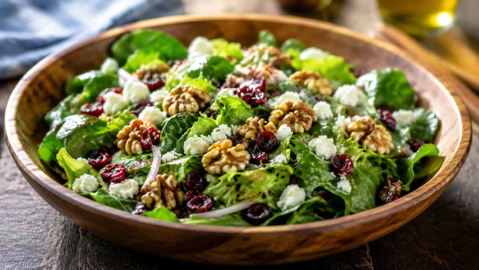 a colorful cranberry walnut salad in a wide wooden bowl in a wide horizontal shot, glossy greens, jeweled cranberries and crumbled cheese, bright natural light, shallow depth of field, landscape orientation