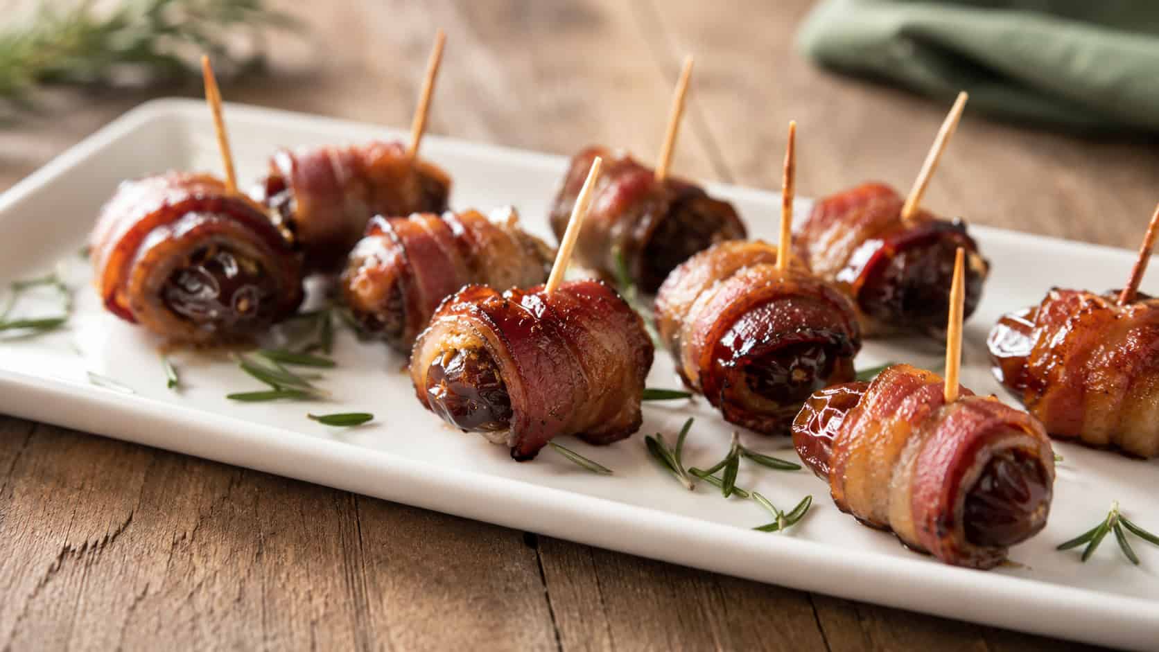 crisp bacon-wrapped dates on a small white platter in a wide horizontal shot, toothpicks in each, scattered rosemary, warm natural light, shallow depth of field, landscape orientation